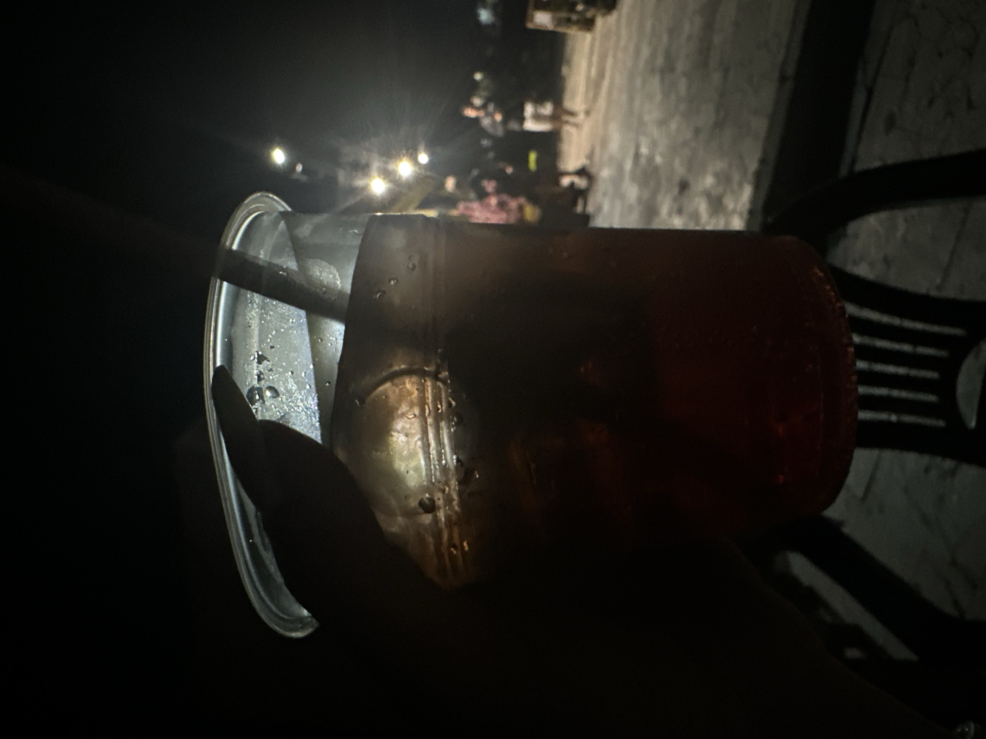 A person holds a clear plastic cup filled with a dark liquid and ice, obscuring a portion of the foreground. The cup is condensation-covered, and a straw is visible within. The background is dimly lit, suggesting nighttime. Several indistinct figures are visible in the distance, appearing to walk along a paved area. Streetlights are present, casting a diffused glow. The scene is set in Galiche, Bulgaria.