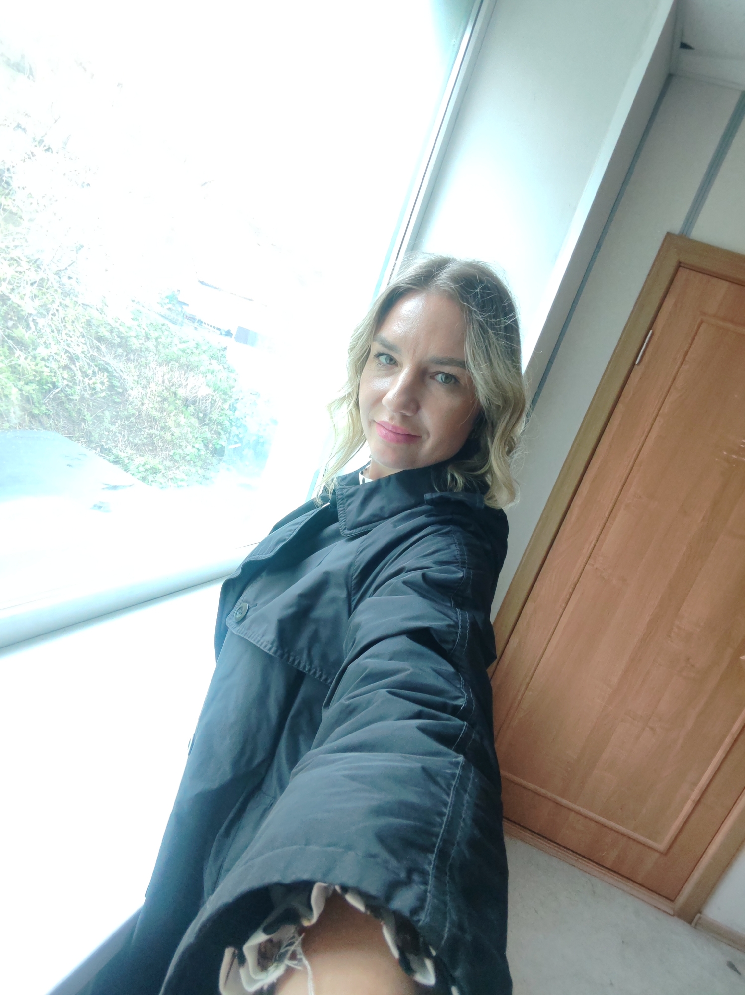A woman with blonde hair is posing for a selfie in what appears to be an indoor setting. She is wearing a black jacket. Behind her is a large window with trees visible outside. To her right, there is a wooden door. The photo does not provide enough information to confirm the location as Petropavlovsk-Kamchatsky, Russia.