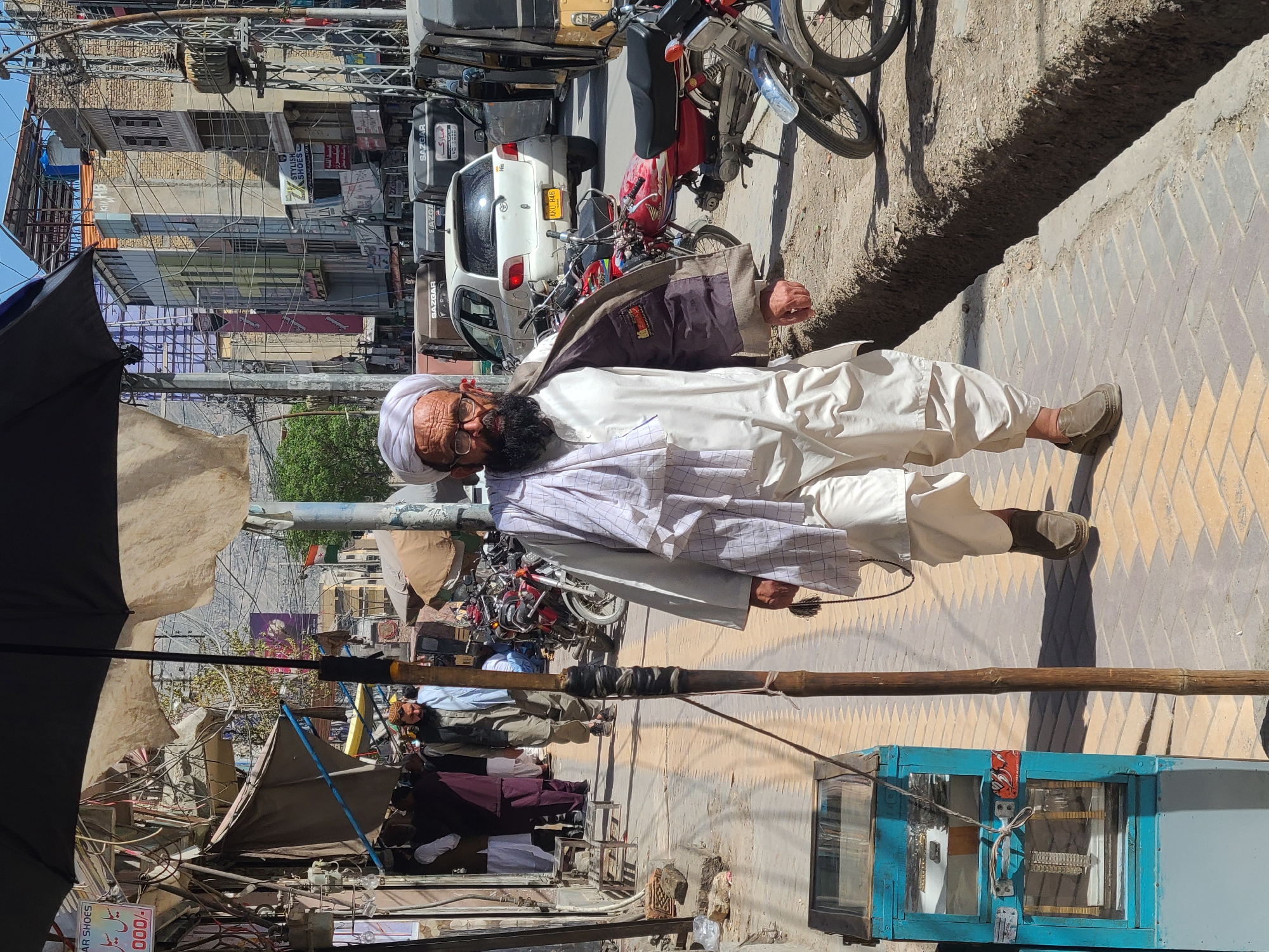 A man wearing traditional Pakistani clothing walks along a sidewalk in Quetta City Tehsil, Pakistan. The street is crowded with cars, motorcycles, and vendors, while buildings are visible in the background. The man is holding a walking stick and has a beard.