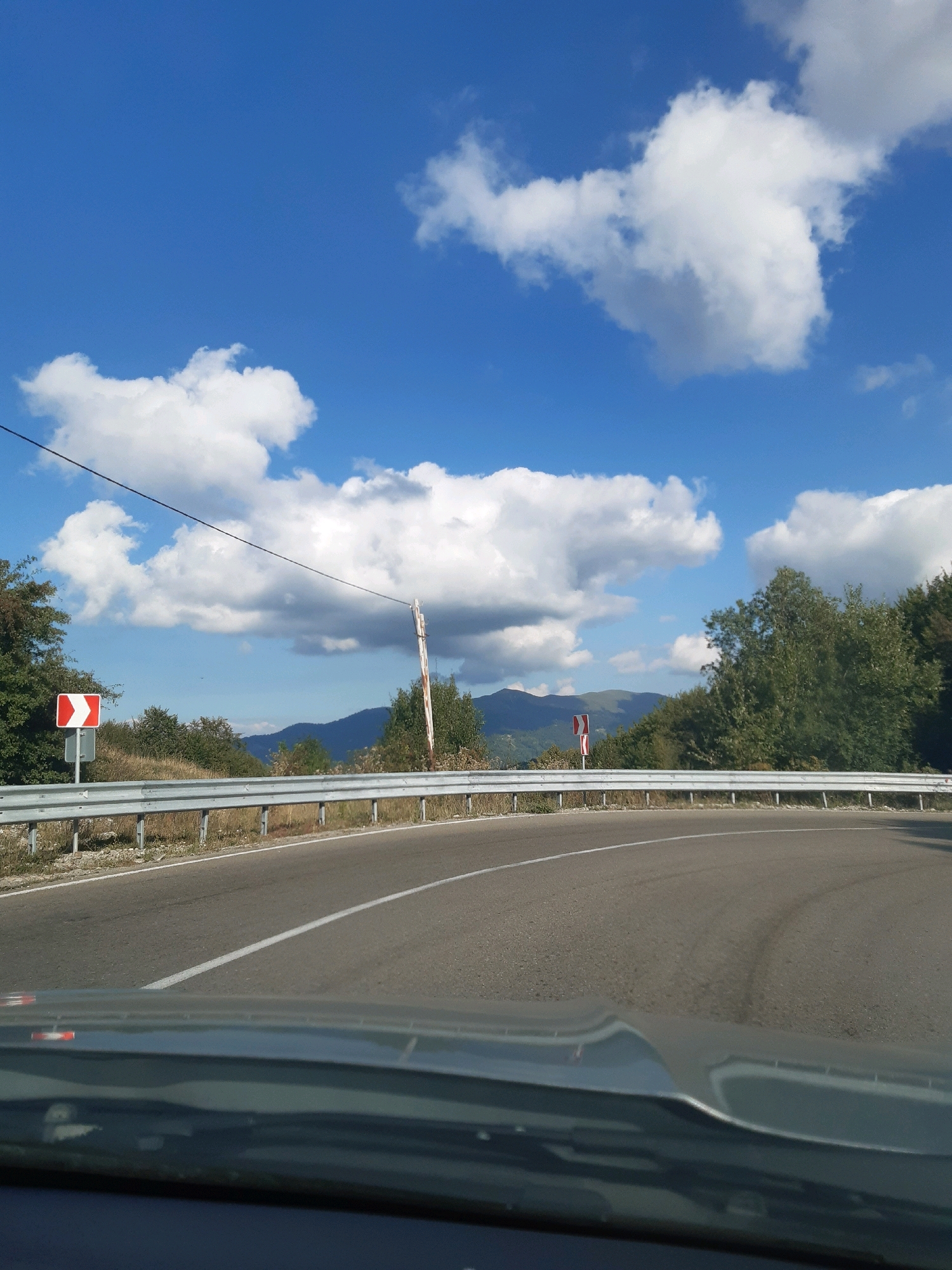 The image depicts a curved road with a guardrail on the left side. A white line marks the center of the road. On the left, a traffic sign with red and white chevrons indicates a curve. Behind the guardrail, dry grass and some trees are visible. In the background, rolling hills covered with trees under a bright blue sky with scattered white clouds are seen. The viewpoint appears to be from inside a vehicle, looking forward. No people are visible. The location is specified as the city of Verona, Georgia.