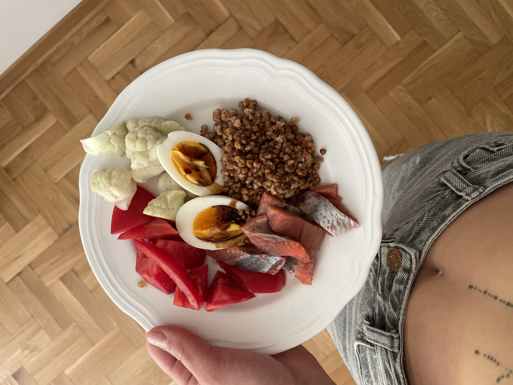 An overhead shot depicts a person holding a white plate of food indoors. The plate contains cauliflower florets, sliced red peppers, two halved hard-boiled eggs, a mound of buckwheat, and several slices of cured fish. The person is wearing gray denim overalls. A navel is visible above the waistband of the overalls, along with a tattoo. The background shows a wooden parquet floor. No location details or people are visible other than what is described. There is no information available about Boreti, Montenegro.