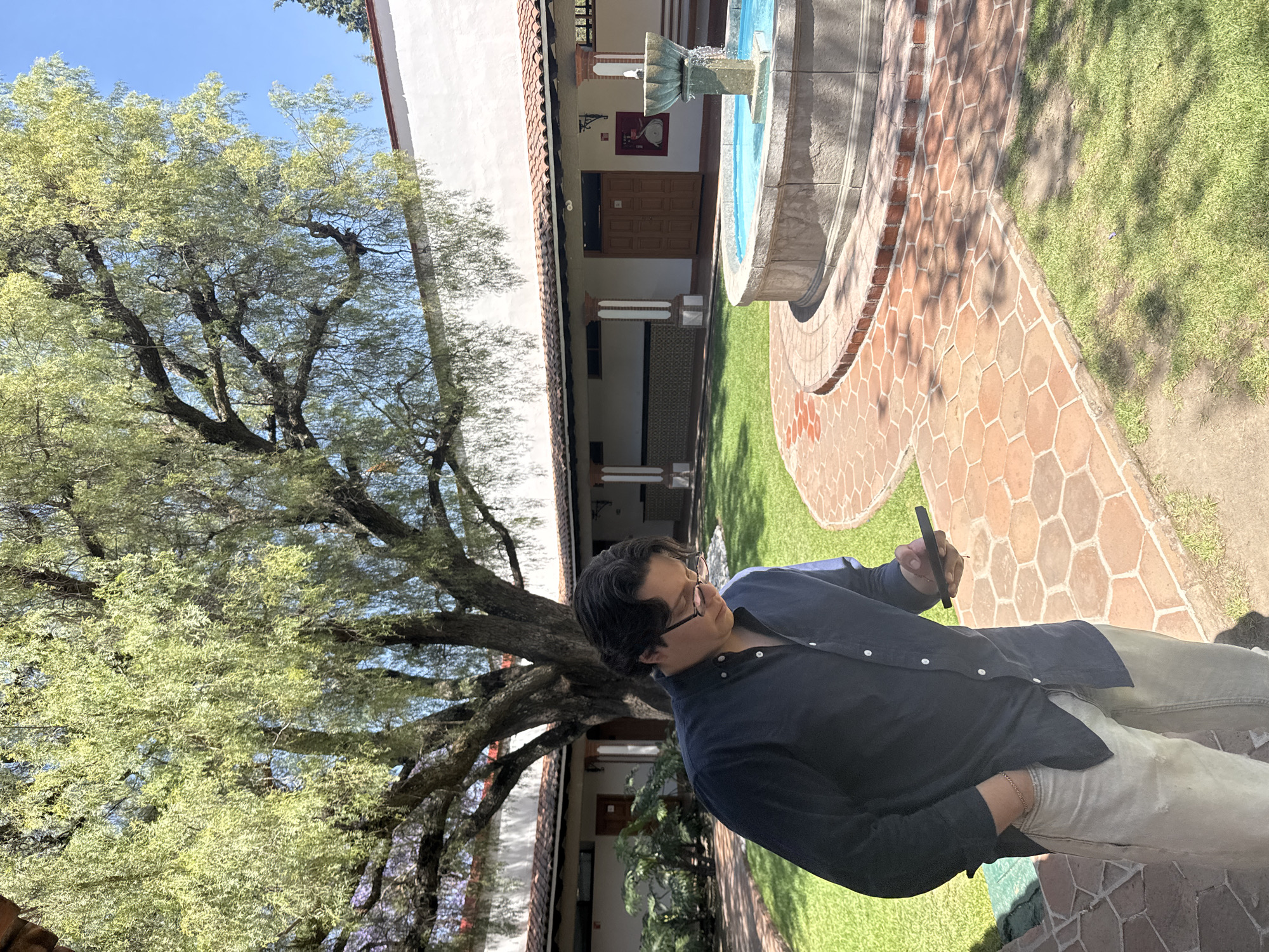 An outdoor courtyard in San Andrés Cholula, Mexico, is depicted under bright daylight. In the foreground, slightly left of center, a male subject with dark hair and glasses stands, looking down at a black smartphone held in his right hand. His left hand is placed in the pocket of his light-colored pants. He wears a dark blue collared button-up shirt with sleeves slightly rolled. The courtyard features a large tree with light green, drooping leaves dominating the left and upper center, casting shadows on the ground. A curved pathway constructed of reddish-brown hexagonal pavers runs from the lower right towards the center, encircling a multi-tiered stone fountain. The fountain, positioned on the right, contains visible blue water actively flowing from its upper tier. Green grass covers the areas between the pathway and the courtyard's perimeter. In the background, white stucco buildings with terracotta-tiled roofs and support columns form a covered walkway. Brown wooden double doors are visible on the building's facade, along with a red wall-mounted object next to one set of doors. A clear blue sky is visible above the tree canopy and building line.