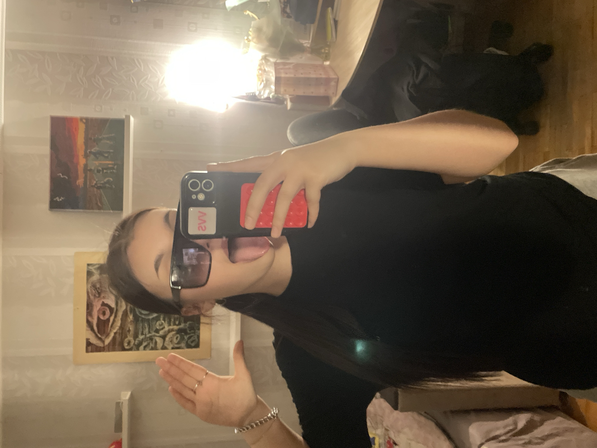 An indoor mirror selfie features a young person posing playfully. The subject has long, dark hair and is wearing a black top and dark-tinted rectangular sunglasses. They are holding a black smartphone with a red textured pop socket or grip in their right hand, using it to capture the image. A sticker with the text "VV" is visible on the back of the phone. The person is sticking out their tongue and has their left hand raised, adorned with rings on some fingers and a silver-colored chain bracelet on their wrist.
The background, reflected in the mirror, reveals a room with light-colored wallpaper featuring a subtle leaf-like pattern. Two framed artworks are hanging on the wall. The upper painting depicts a landscape with a dark ground and an orange to dark sky, possibly showing figures or structures. The lower painting, framed in light wood, features an abstract or stylized organic form. A very bright artificial light source, likely a ceiling fixture, causes a strong glare in the upper right corner of the reflection. Other indistinct elements of a room, including what might be furniture and general household items, are visible in the background. The scene suggests an evening or night setting due to the reliance on artificial lighting. The location is Murino, Russia.