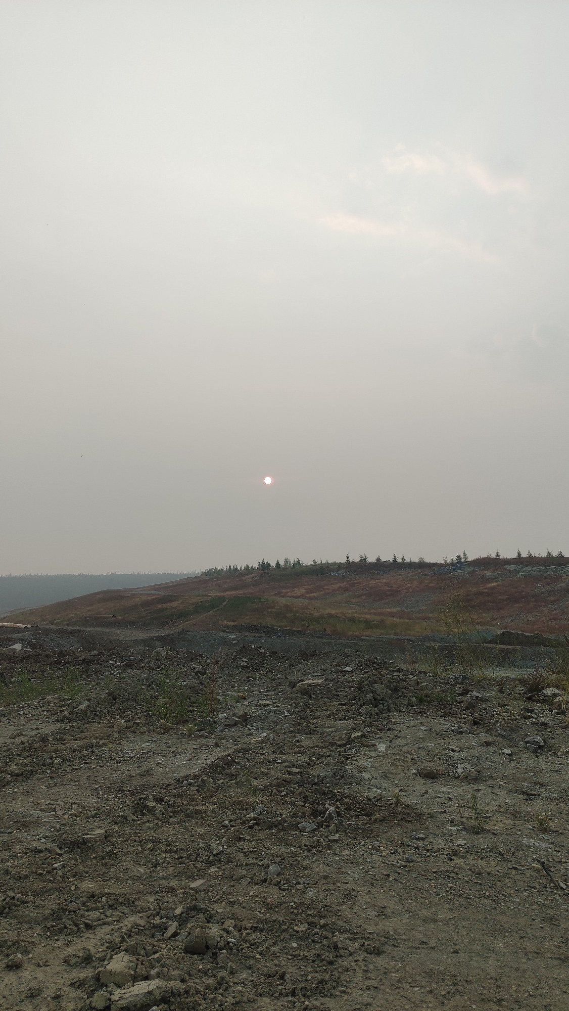 The image depicts a landscape with a hazy sky, likely due to smoke or dust, obscuring the sun which appears as a small, reddish-orange orb. The foreground is a rough, uneven terrain covered in dirt, rocks, and gravel. In the mid-ground, rolling hills are covered in dry vegetation and sparse trees, with a line of evergreens along the crest of the furthest hill. The overall impression is of an industrial or mining area, given the rugged terrain and lack of greenery. The scene is devoid of any people or obvious signs of activity. The location, Mirny, Russia, is known for its diamond mines, which could explain the disturbed earth and barren landscape.