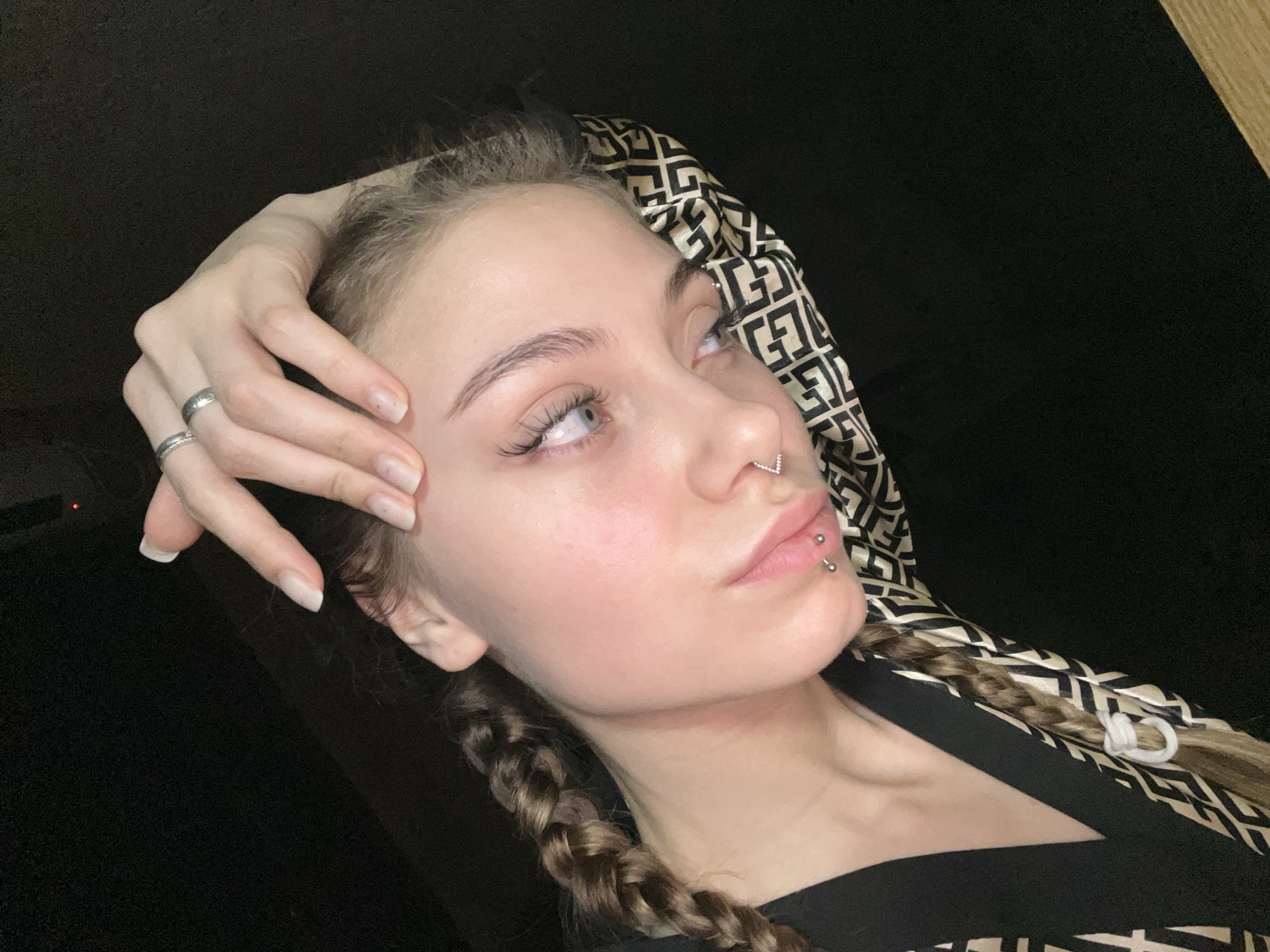 A young woman is depicted in a medium shot, facing upwards and slightly to the right. She has light skin, blue eyes, and blonde hair styled in braids. Her left hand is positioned near her temple, with two rings visible on her fingers. She has multiple piercings, including one in her nose and two on her lower lip. She is wearing a patterned headscarf and a dark-colored top. The background is black. It is not possible to determine a specific location in the city of Фоки, Russia from the image.