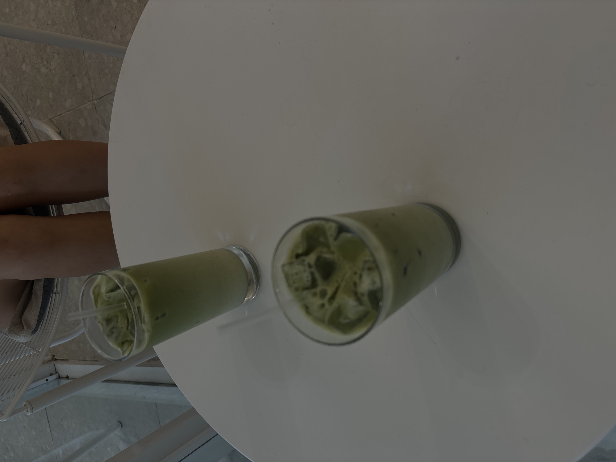 Two glasses of iced matcha latte sit on a white table. The glasses are tall and clear, filled with a pale green liquid and ice cubes. Straws are present in both glasses. In the background, a person's legs are visible, wearing shorts. The person is seated on a white chair with a woven seat. The flooring appears to be tiled. The scene is set indoors, likely in a cafe or restaurant. The lighting is soft and diffused. The image does not provide any information that would suggest its location in San Juan, United States of America.