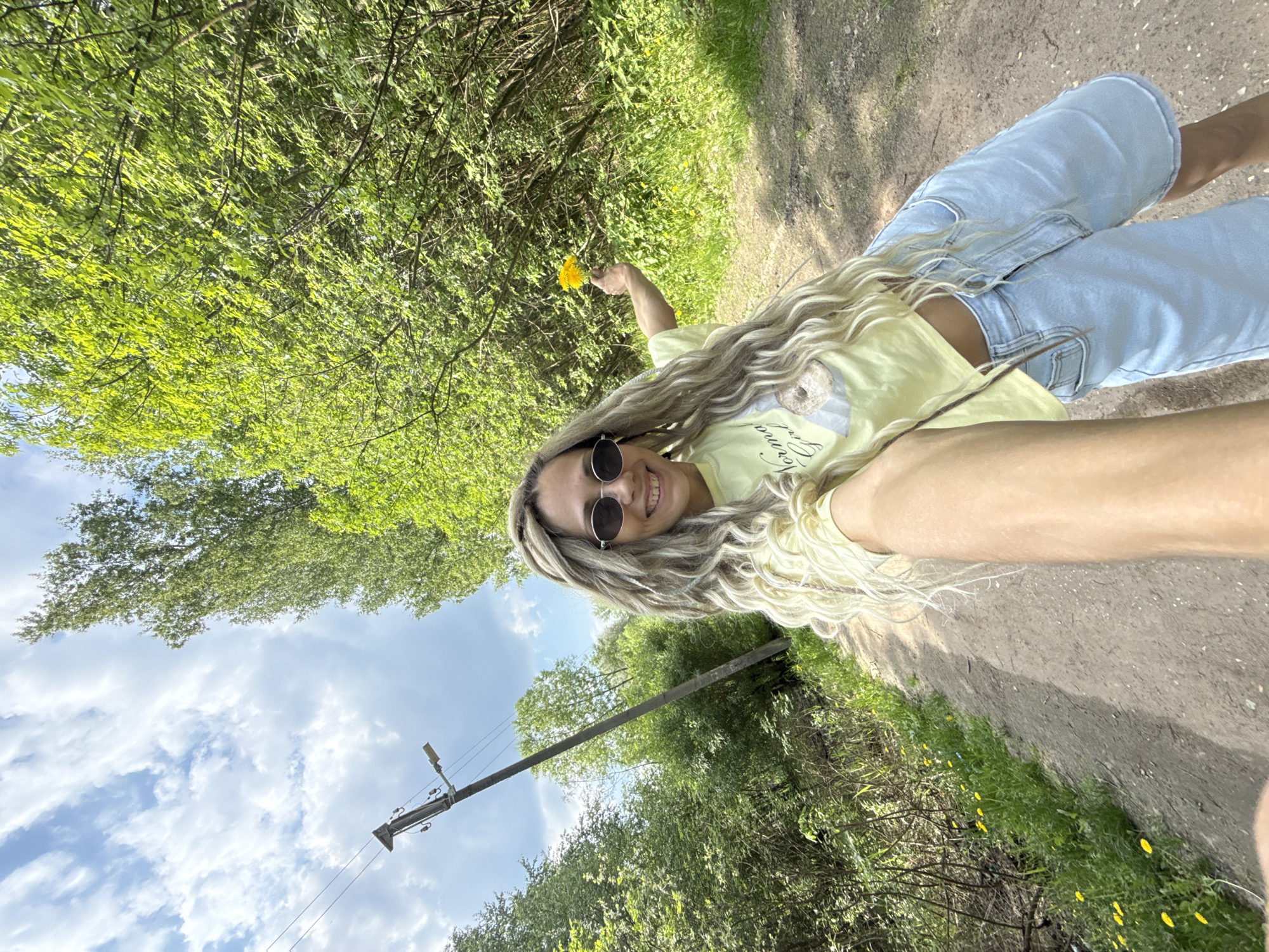 A young woman with long blonde hair, wearing a yellow t-shirt, light blue jeans, and sunglasses, is holding a dandelion flower and smiling for a selfie on a dirt path. The background includes trees, foliage, a utility pole, and a partly cloudy sky. The location is likely Rusanovo, Russia.