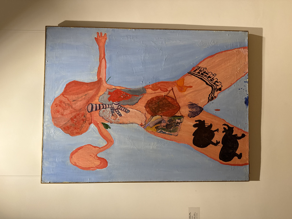 An abstract painting shows a reclining, nude figure on a light blue background. The figure is primarily pink, with internal organs depicted in red and brown. The head is misshapen with long, twisting hair. One arm is raised. Dark silhouettes of dancing figures appear near the hip. A striped object is near the chest. The figure's thigh features a patterned band. The painting is framed.