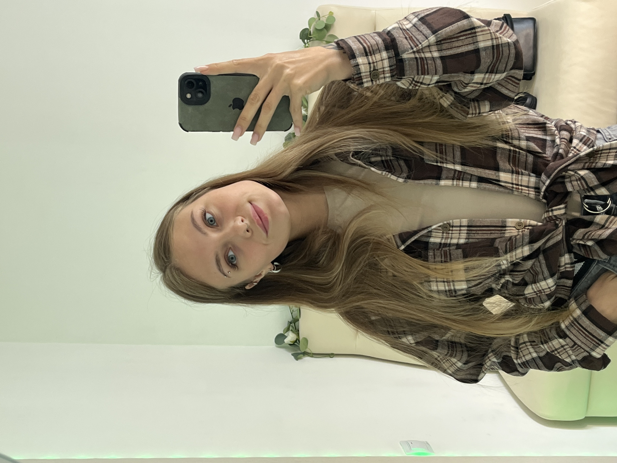 A young woman with long blonde hair, wearing a plaid shirt and a beige t-shirt, takes a selfie in a brightly lit room. She is holding a dark-colored smartphone in her right hand. The background shows a plain wall and a chair with some greenery. There are no identifiable landmarks or clues that would indicate the location as Фоки, Russia.