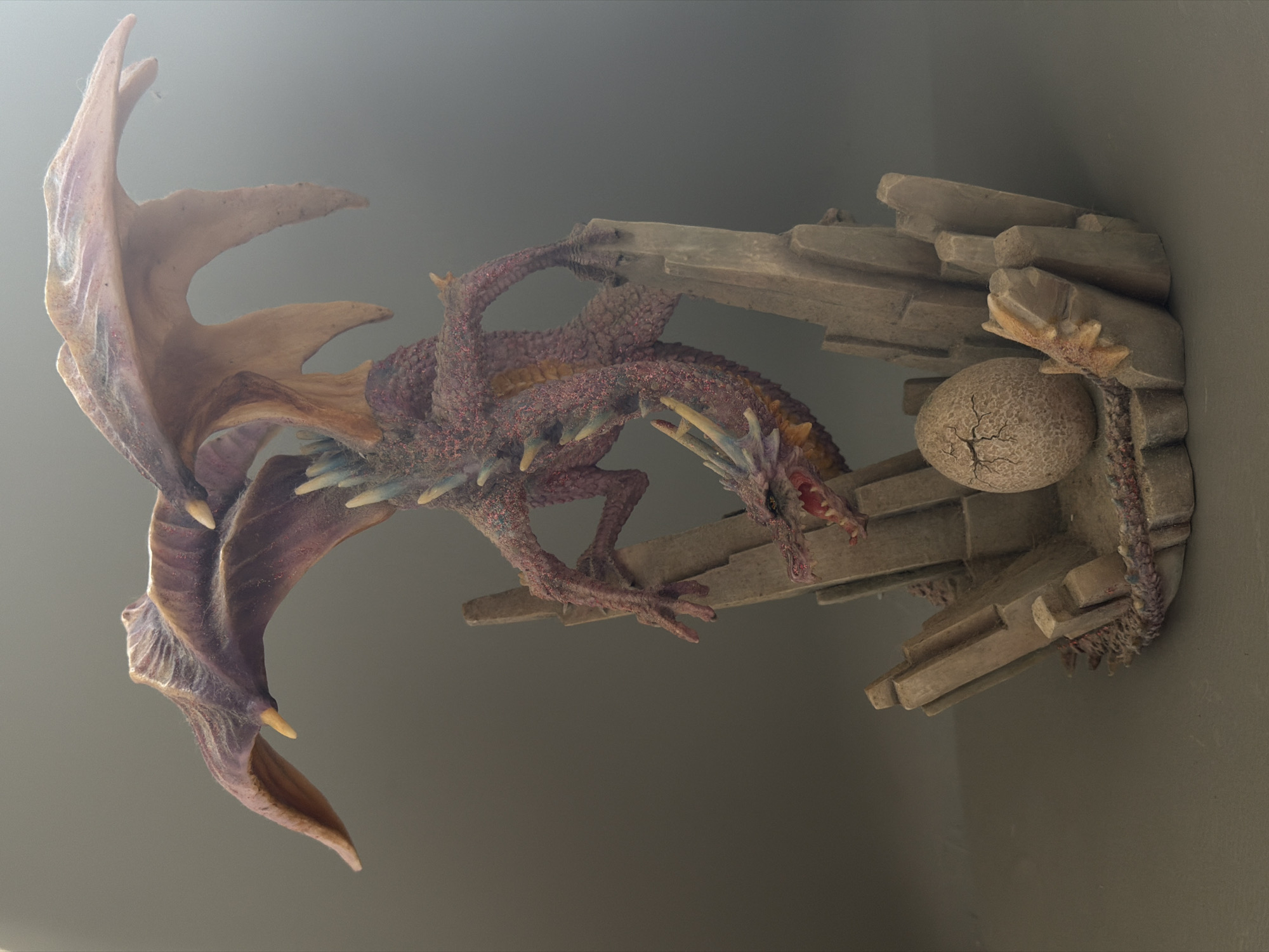 This media file shows a detailed sculpture of a mythical creature, likely a dragon, perched on a rocky outcrop. The dragon is predominantly colored in shades of reddish-purple, with iridescent speckles, and has large, translucent wings that are a lighter, sandy brown color with purple undertones. Its mouth is open, revealing sharp teeth and a pink interior. The dragon is positioned as if it is either landing or taking off from the rocky structure, with its claws gripping the stone.
The rocky outcrop appears to be made of a light grey material, textured to resemble natural rock formations with layered, angular pieces. A notable detail on the outcrop is a single, cracked egg, also made of the same stone-like material, resting near the dragon. The overall scene is staged against a neutral, softly lit grey background, suggesting an indoor display or a studio shot. The lighting casts subtle shadows, highlighting the textures and details of both the dragon and its rocky perch. The focus is on the artistry of the sculpture, emphasizing the intricate scales of the dragon, the delicate structure of its wings, and the realistic weathering of the rock and egg. No text is visible in the image.