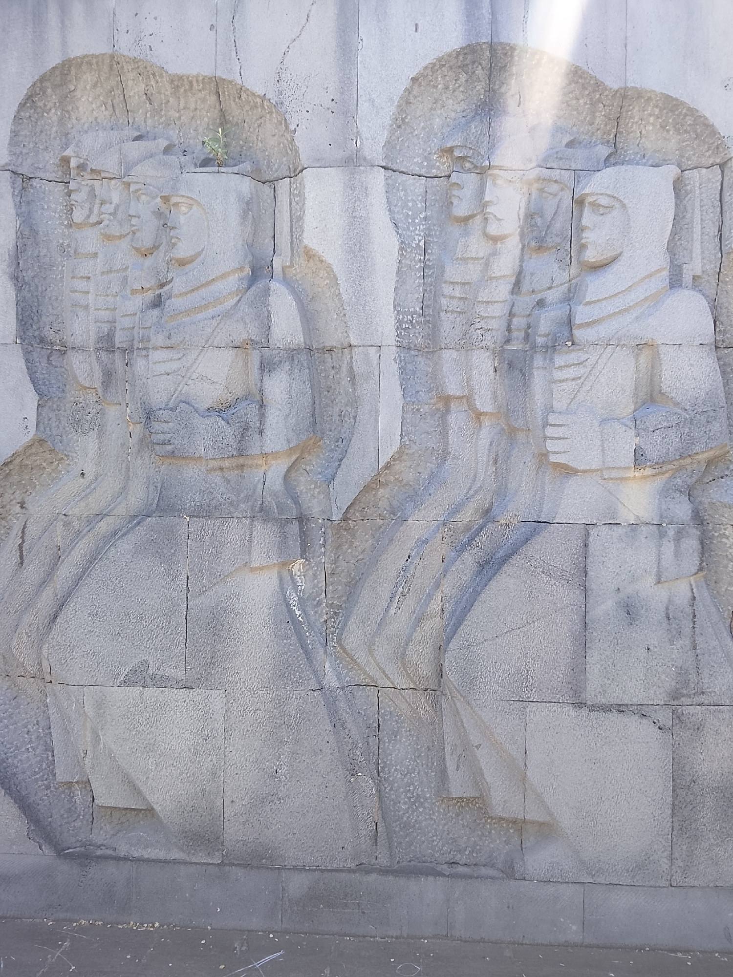 A weathered stone relief sculpture is depicted on a light-colored wall under bright daylight. The sculpture features multiple stylized human figures, possibly soldiers or historical individuals, carved in profile and arranged in rows. These figures appear to be facing towards the right. They wear distinct headwear and garments, with some figures showing detailed facial features like moustaches, while others are more obscured. On the left side, a small green plant grows out of a crack in the stone above the figures' heads. On the right, a prominent figure in a hood-like head covering holds an object, possibly a book or tablet, in their hands. The figures are depicted from the waist up, creating a sense of a unified procession or group. The stone blocks of the wall and the sculpture show signs of age, including cracks and a textured surface. Strong sunlight casts defined shadows and highlights across the relief, emphasizing its three-dimensional quality. The base of the wall and the ground below are visible, suggesting an outdoor setting. No text is visible in the image.