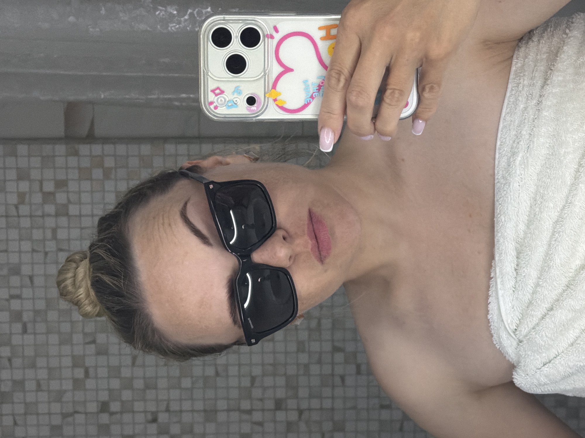 A woman with blonde hair pulled back in a bun and wearing black sunglasses is taking a selfie in a bathroom. She is holding a phone with a white case decorated with a pink heart and cartoon images. She is partially wrapped in a white towel. The background shows gray and white tiled walls. The location is presumed to be a bathroom, although a more specific location in the city of Votkinsk, Russia, cannot be determined from the image.