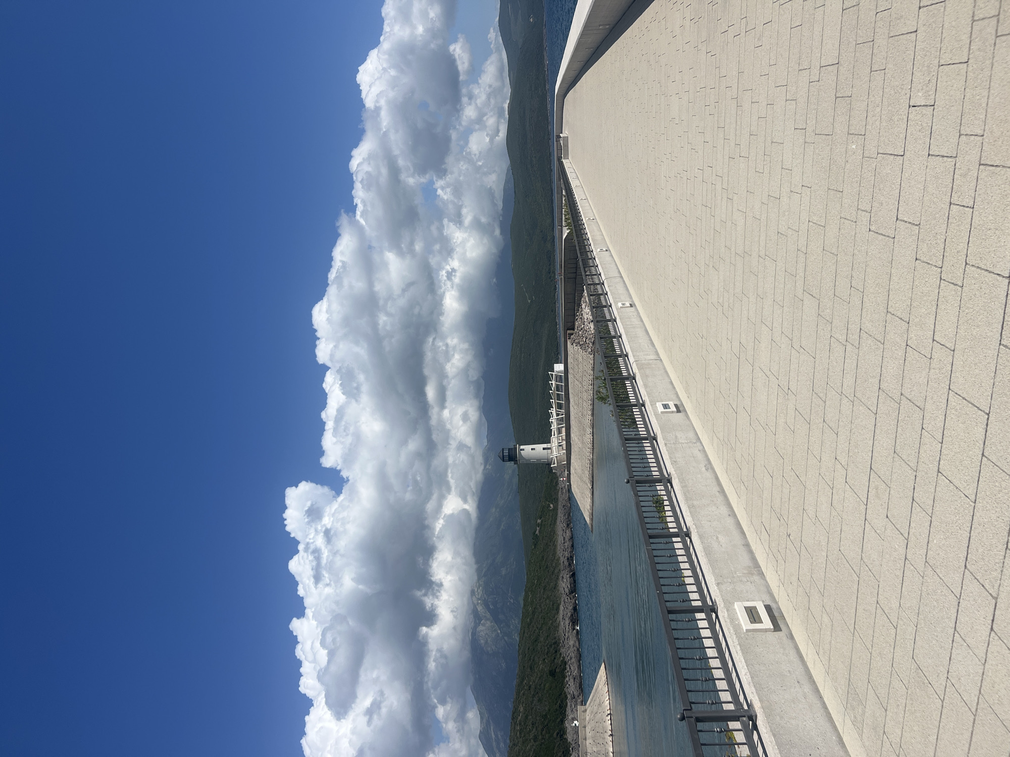 The image shows a waterfront scene in Radovići, Montenegro. A large, white, brick-paved promenade extends along the right side of the frame. A dark metal railing borders the promenade along the water's edge. Below the promenade is the shoreline, with a small lighthouse structure. In the background are mountainous terrain and a blue sky partially obscured by large, fluffy white clouds.