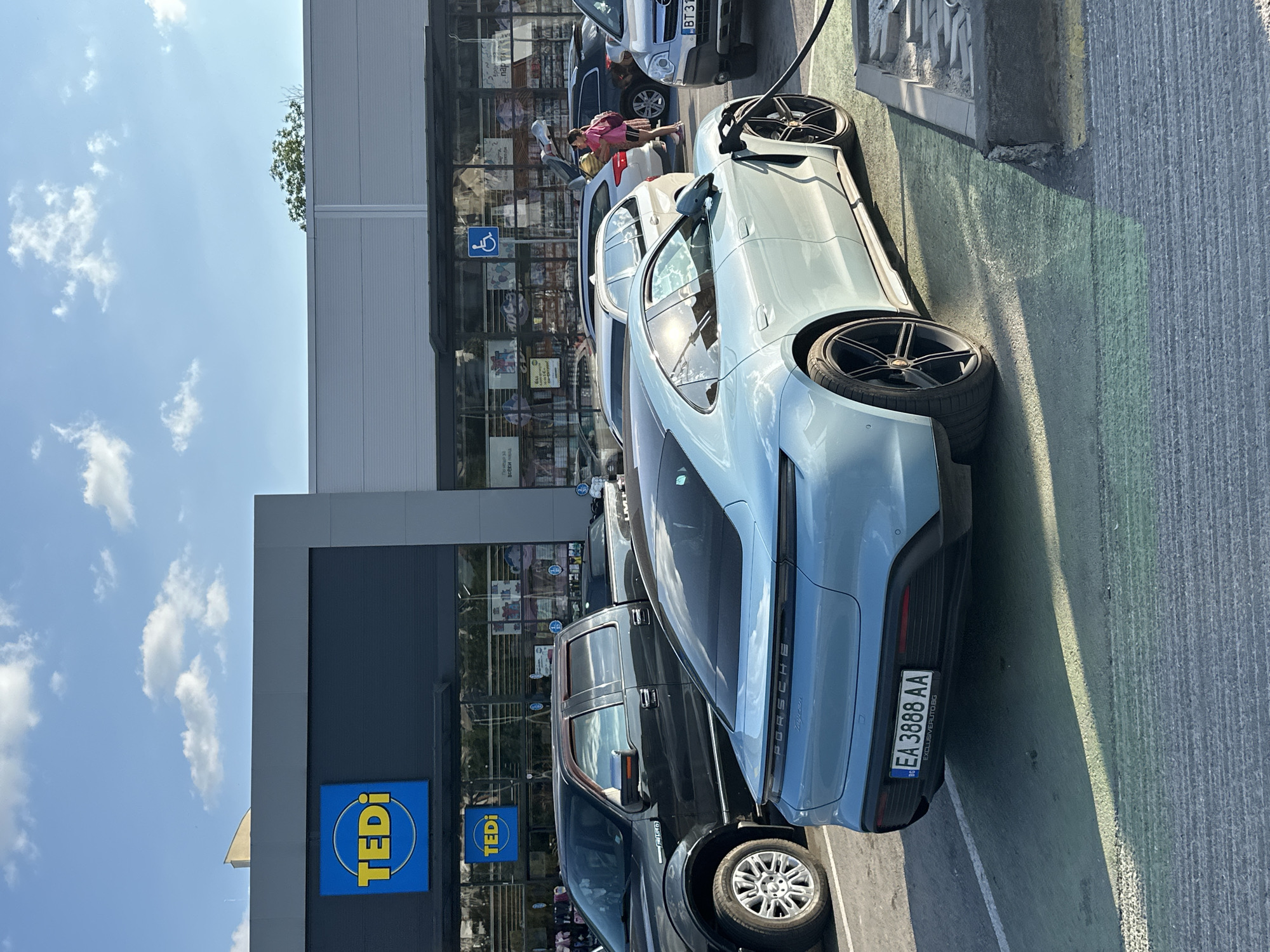 The image shows a bright, sunny day in Veliko Tarnovo, Bulgaria. The primary subject is a light blue Porsche Taycan parked at a charging station, its charging cable plugged in. Beside it, a dark pickup truck is also parked, and further back, several other cars are visible, including a silver SUV. In the background, a building with large glass windows is adorned with signs, including a blue and yellow "TEDI" logo. A person in a pink shirt is visible near the entrance of the building. The scene appears to be a parking lot outside a commercial establishment, possibly a shopping center or a store. The sky is clear with scattered white clouds. The overall impression is one of a typical day in a Bulgarian town, with vehicles and people going about their business. The license plate of the Porsche is visible, reading "EA 3888 AA".