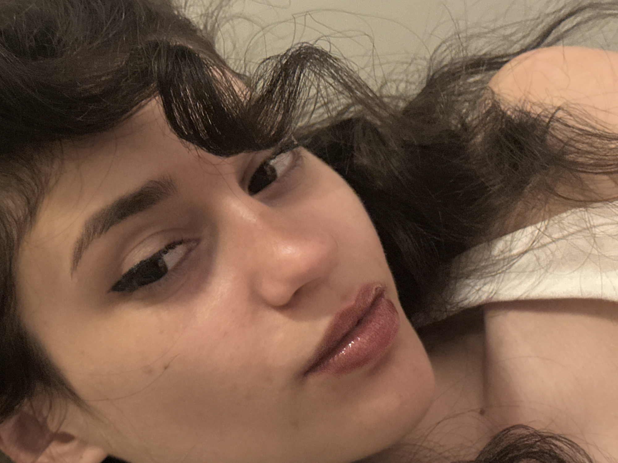 The image is a close-up, low-angle, headshot of a young woman with dark, curly hair and light skin. She is wearing a white tank top and has winged eyeliner and glossy, brownish-red lipstick. Her head is tilted to the left, and her gaze is directed towards the viewer's left. The lighting is warm and soft, casting shadows that accentuate the contours of her face. The background is a plain, neutral color, possibly a wall. There is no information or visual cues in the image to suggest a specific location in the city of Sofia, Bulgaria.