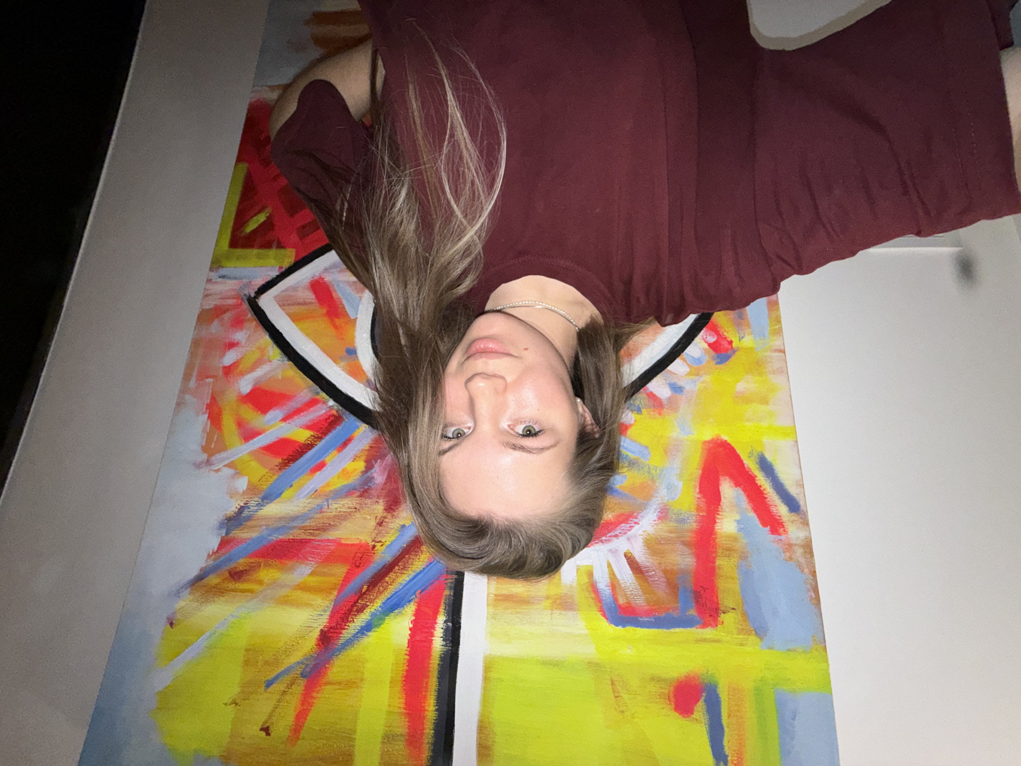 A young woman is posing with a large abstract painting. The painting features a yellow and white background with overlaid strokes of red and blue paint, forming geometric shapes. The woman is wearing a maroon t-shirt and a silver necklace. The background behind the painting is a plain white wall. The photo is taken indoors and oriented upside down. It is not possible to know its location in Vozdovac Urban Municipality, Serbia.