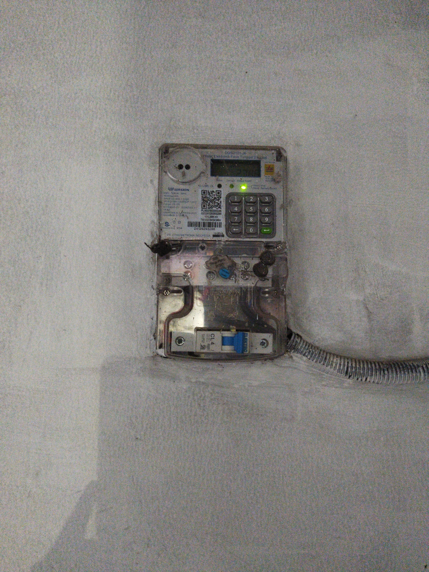 The image shows an electric meter mounted on a textured, light-colored wall. The meter, which appears to be a smart meter, is encased in a clear plastic housing. It has a digital display, a keypad with numbers 0-9 and an enter button, and a green light indicating it is on. Labels on the meter include "DDS D101-JS", "Listrik Fase Tunggal 2 Kawat", and a QR code. A circuit breaker is visible below the meter, labeled "CL4" and "ELECTRO". A corrugated pipe with electrical wiring emerges from the bottom right of the meter enclosure and runs along the wall. The surrounding wall is unevenly textured and appears to be painted or plastered. The lighting suggests an indoor or covered outdoor environment. The overall scene is utilitarian and functional, depicting standard electrical infrastructure.