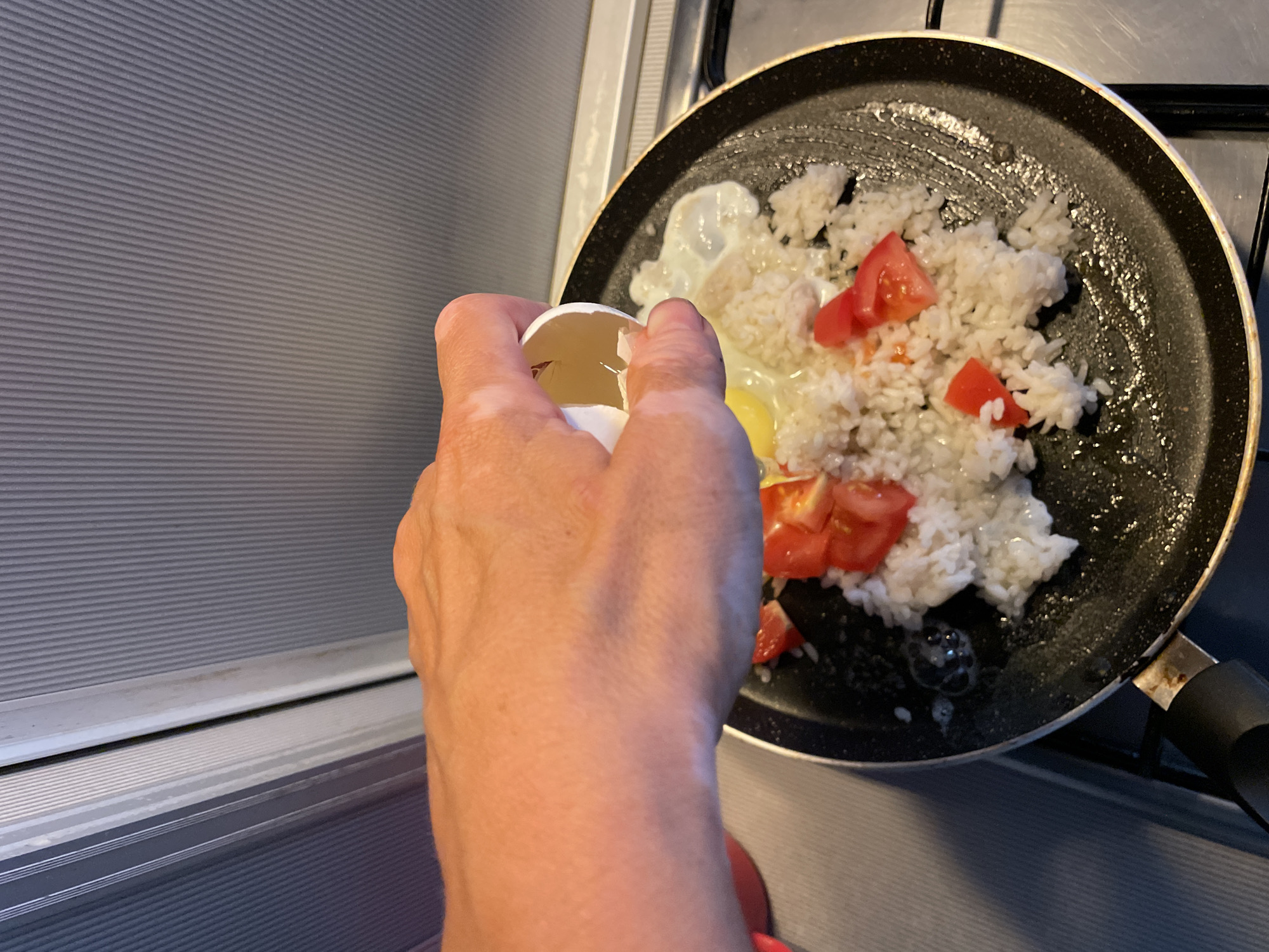 An overhead shot captures a hand cracking an egg into a frying pan containing rice and chopped tomatoes. The scene appears to be a kitchen counter in Sochi, Russia, with a stovetop visible. The pan is black and circular, with oil sizzling around the food. The hand is light-skinned, with some sunspots. The background consists of a gray, ribbed wall.