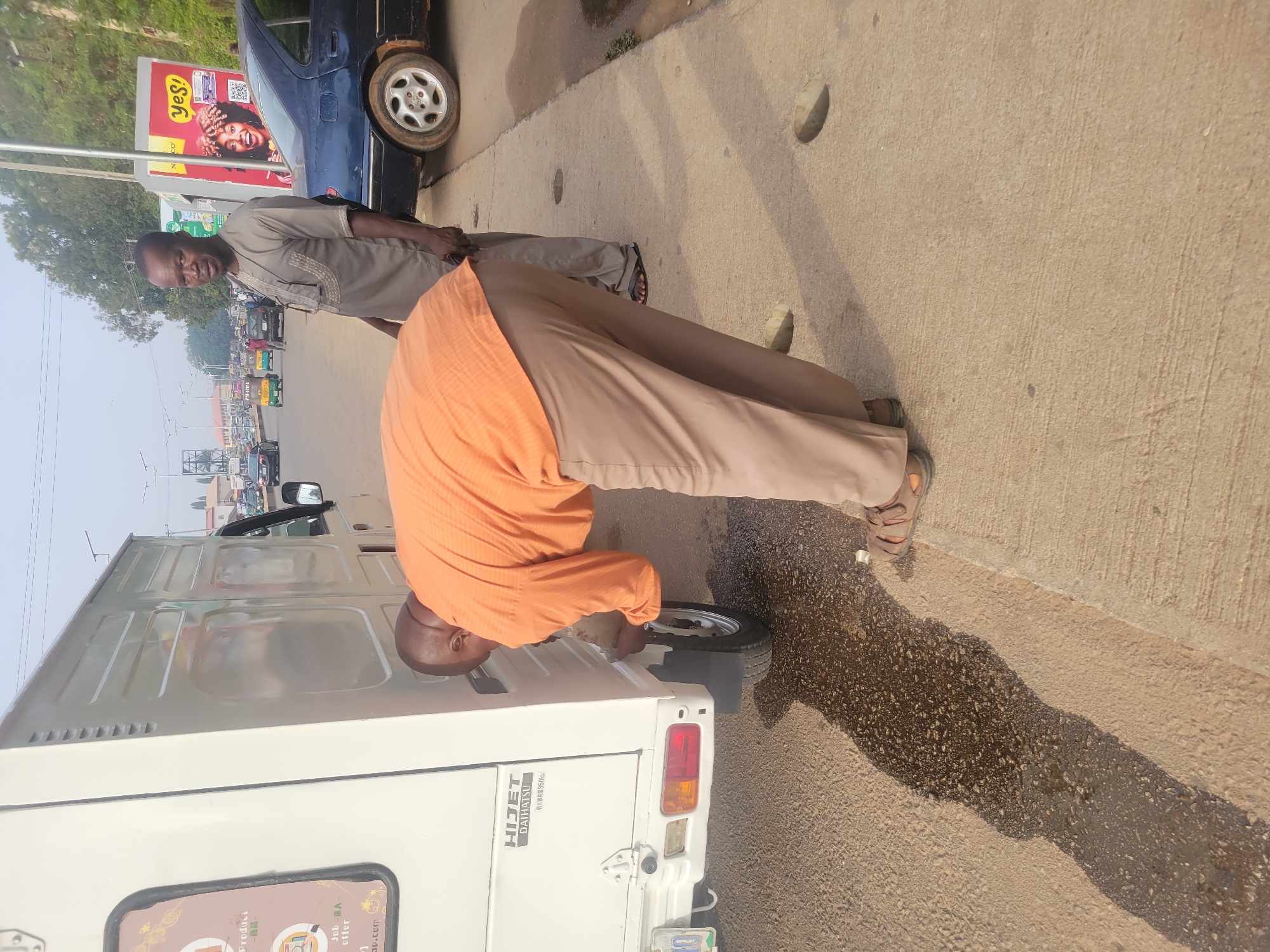 Two men are standing near a white Hijet Daihatsu truck on a roadside. The man closer to the truck is wearing an orange shirt and khaki pants, and is inspecting the rear tire. The other man is wearing a beige shirt and khaki pants. A dark blue car is parked nearby. A billboard with the word "Yes!" is visible in the background, along with a street scene with cars and buildings. The location appears to be in the city of Jos, Nigeria, as indicated by text on the truck.