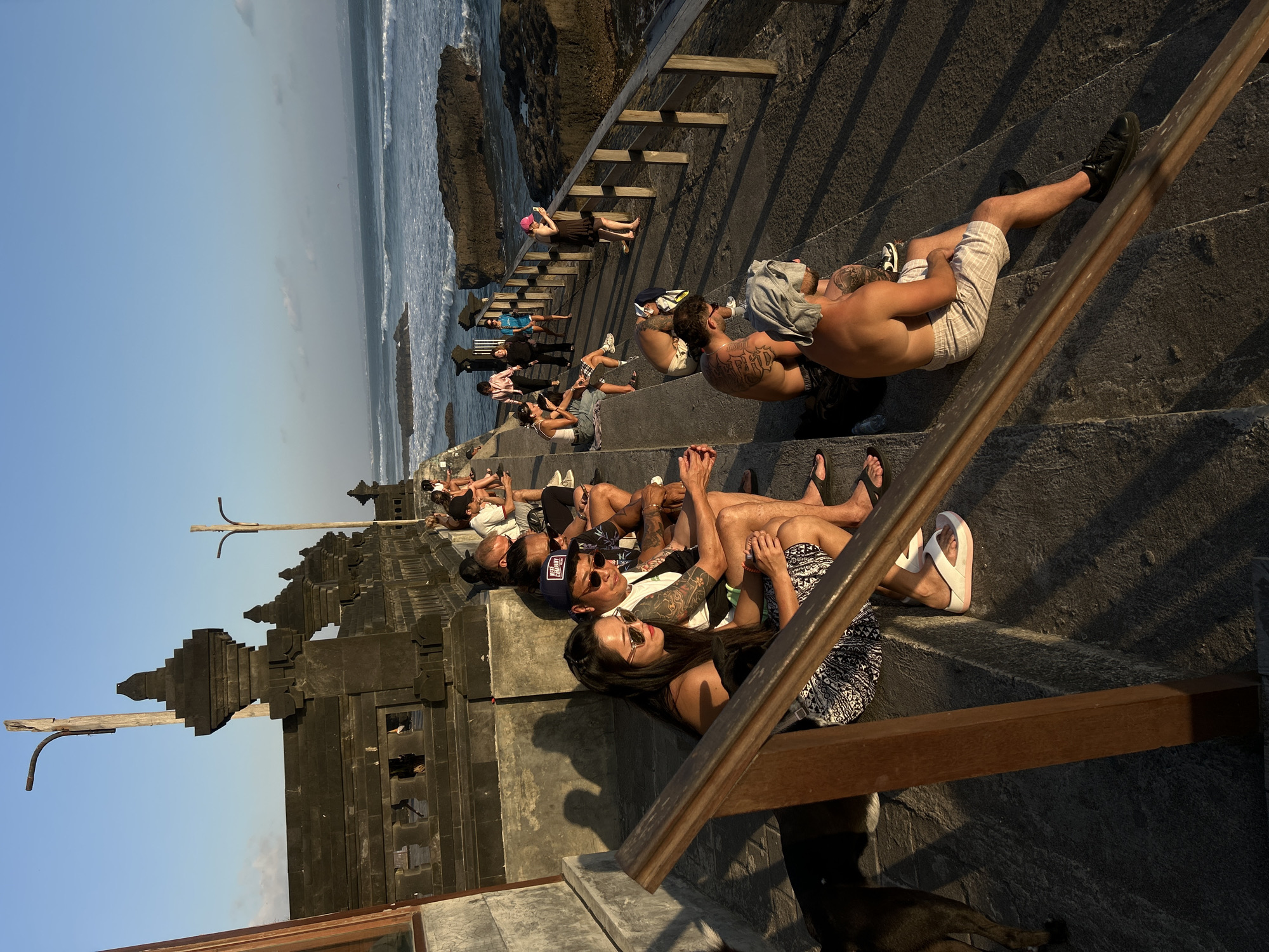 In North Kuta, Indonesia, a group of people are seated on stone steps facing the ocean. The setting appears to be a temple or a similar cultural landmark, indicated by the dark, ornate stone structures in the background and on the left side of the image. The sea is visible in the distance with waves crashing against dark rocks. The sun is low in the sky, casting long shadows and illuminating the scene with warm, golden light. Several individuals are dressed casually, some wearing sunglasses and hats. A black dog is present in the foreground, to the left of the frame. The people are engaged in various activities, including looking at their phones, talking to each other, and simply observing the view. The atmosphere is relaxed and communal, with individuals sharing the space and enjoying the coastal scenery.