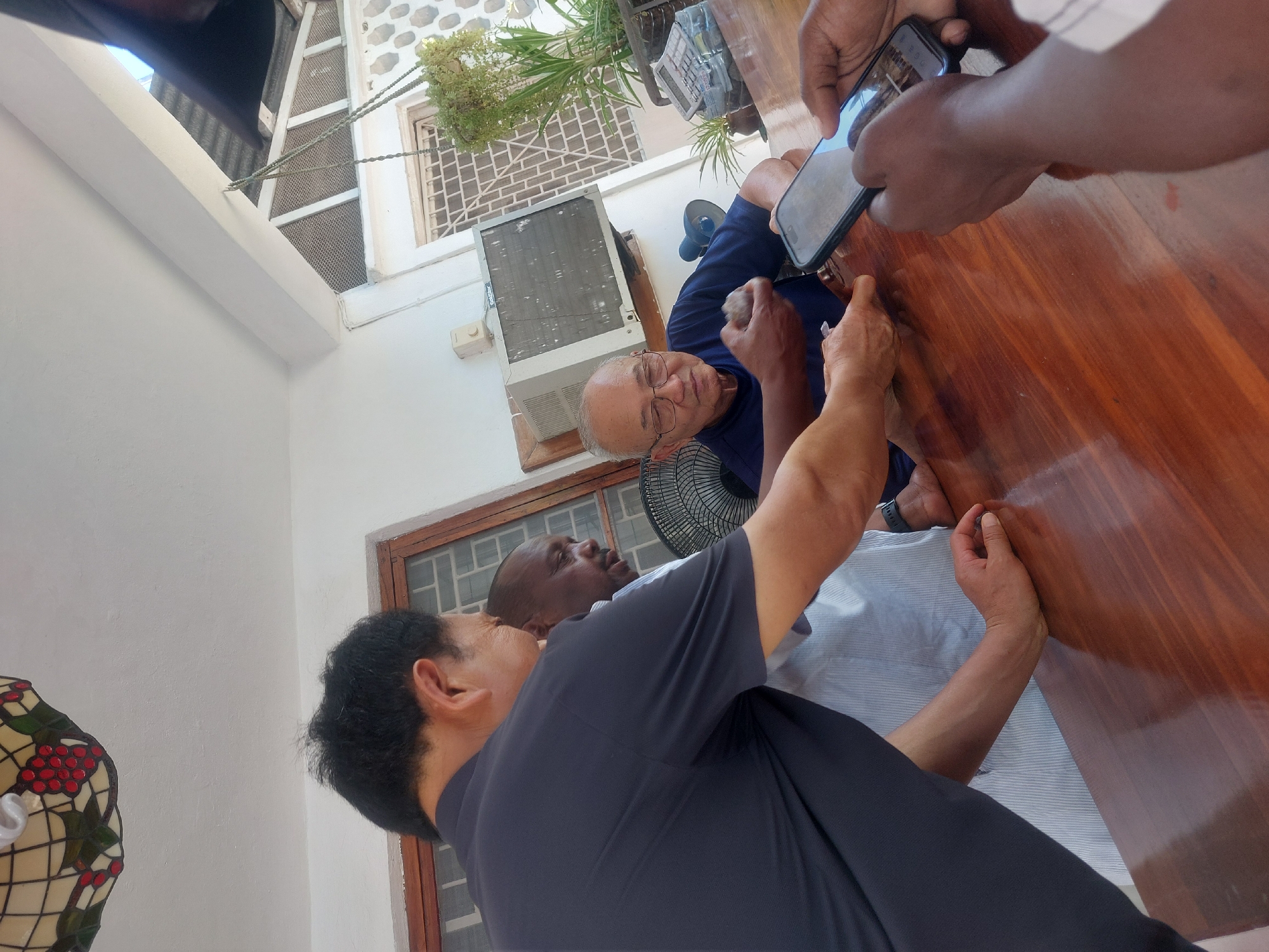 An indoor scene in Dar es-Salaam, Tanzania, captures a gathering of several men around a polished wooden table. The primary focus is an older, light-skinned man with glasses, wearing a blue shirt, seated at the table and looking towards the camera. He appears to be the subject of a recording, as a pair of dark-skinned hands holds a smartphone, pointing it towards him.
To his left, another dark-skinned man in a light-colored striped shirt is partially visible, also looking in the direction of the man in blue. In the foreground, from a slightly overhead perspective, the back of a man with black hair, wearing a dark blue or black shirt, is seen. He is also oriented towards the focal point at the table. Several other hands and arms rest on the table, indicating more people are present but not fully in frame.
The setting appears to be a brightly lit room with white walls. In the background, a wall-mounted air conditioning unit and a small black desk fan are visible. Above the AC unit, a window with grates and some foliage outside suggests a tropical or sub-tropical climate. To the far left, a decorative stained-glass lamp with red berries and green leaves adds a touch of ornamentation. The activity appears to be a casual meeting or social interaction, with one person being filmed or photographed. The time of day seems to be daytime due to the bright ambient light. No discernible text is visible on any objects, clothing, or the phone screen.