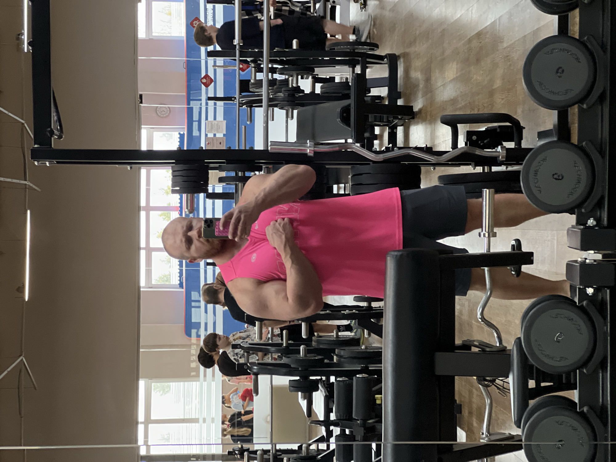 The image depicts a man taking a selfie in a gym setting in what appears to be Chaikovsky, Russia. The gym is equipped with weightlifting machines and free weights. The man is wearing a pink tank top and dark shorts. In the background, other gym-goers are visible, and through the windows, some buildings and trees can be seen. The reflections in the mirror further show the gym environment with different people and equipment.