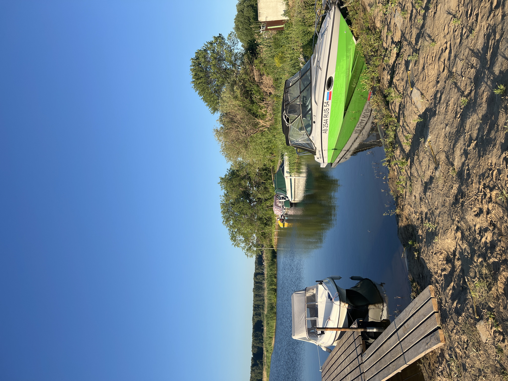The image depicts a serene waterside scene in Novopichugovo, Russia, under a clear blue sky. A wooden pier extends into the calm water on the left, with a white boat moored to it. Further out on the water, several other boats are visible, some covered with tarps. On the right bank, a brightly colored green and white motorboat is partially pulled onto the sandy shore, its reflection shimmering in the water. The background features lush green trees and vegetation, with a distant line of trees bordering the horizon. The overall atmosphere is peaceful and idyllic, suggestive of a recreational area. There are no people visible in the image.