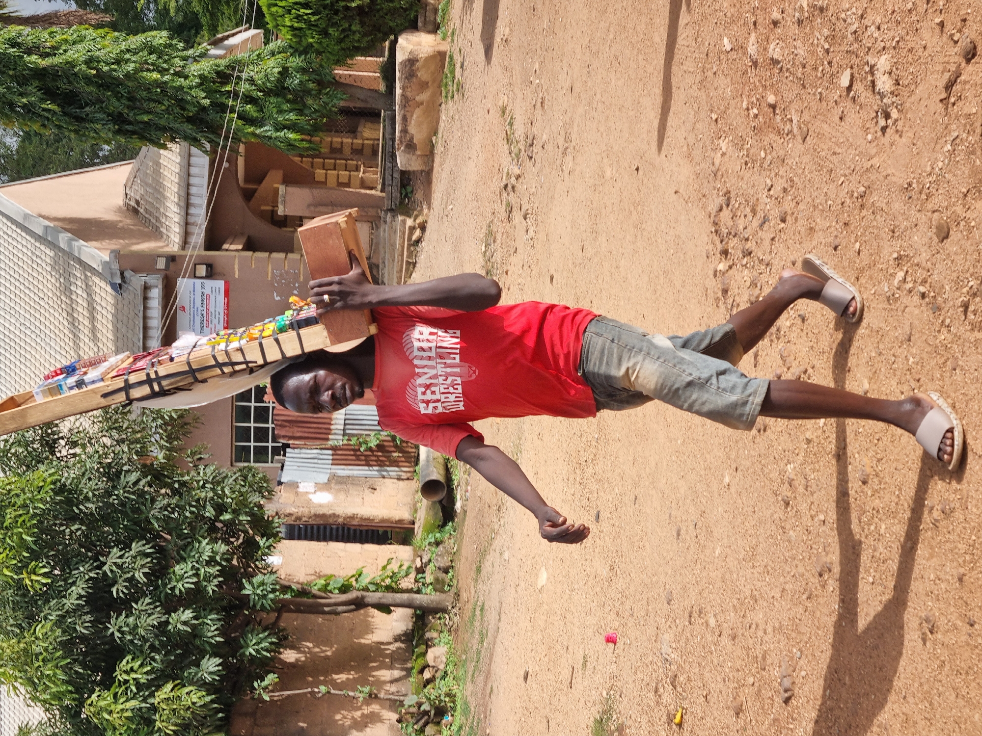 A Black man is walking on a dirt road carrying a wooden object over his shoulder. The man is wearing a red t-shirt with white lettering that says "SENIOR WRESTLING" and denim shorts. He is also wearing light-colored sandals. The wooden object appears to be a display stand with colorful items attached to it. In the background, there is a light brown building with a tiled roof and green trees. The ground is covered in sand and small stones. The image appears to have been taken in a semi-arid environment, possibly a rural area within the city of Jos, Nigeria.