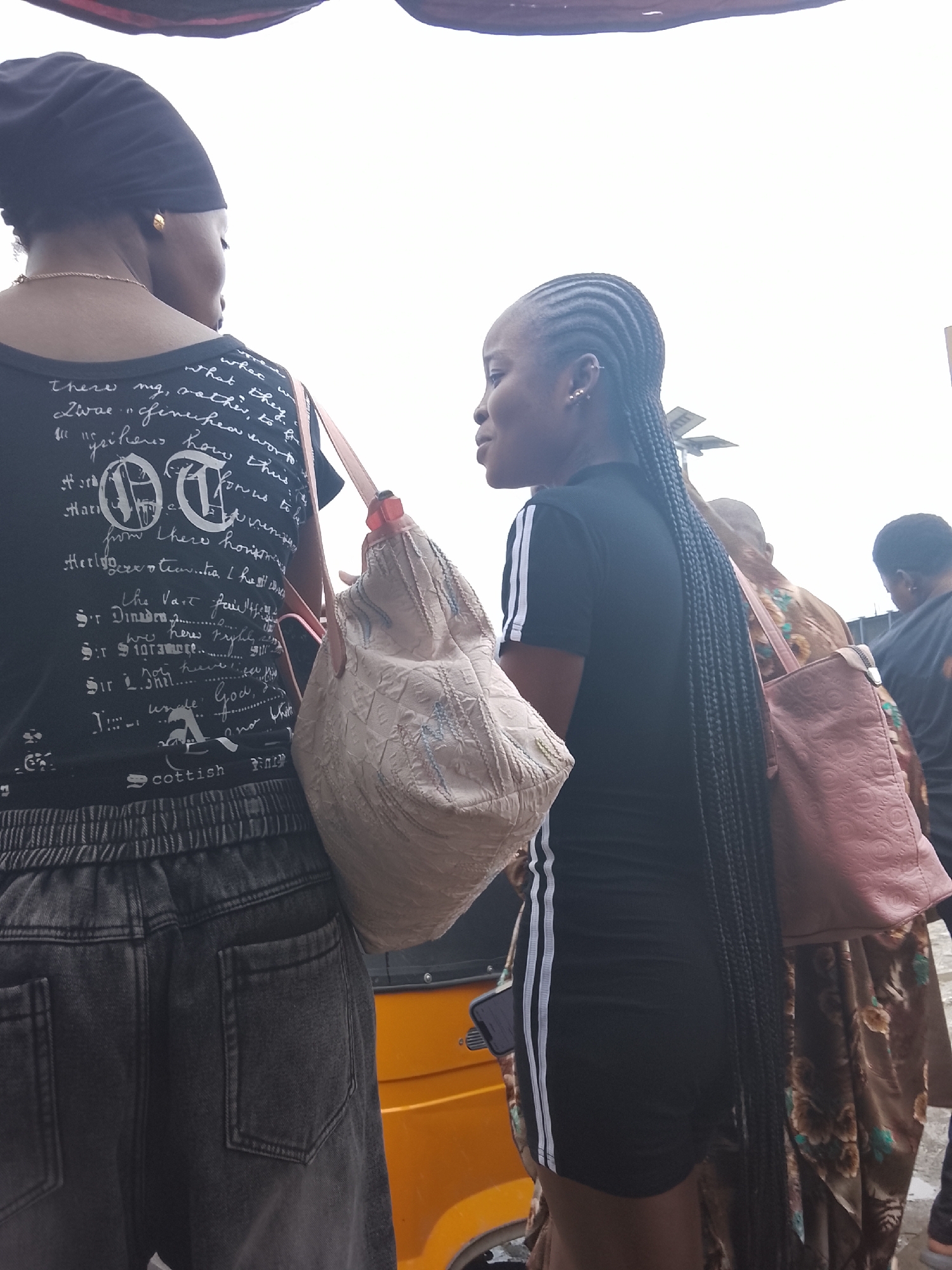 In Lekki, Nigeria, two Black women are standing outdoors, possibly in a marketplace or near a street. The woman on the left wears a black beanie, a black t-shirt with white text and graphics, and dark grey denim pants. She has a gold earring and a gold chain necklace. A light-colored, textured bag hangs from her right shoulder. The woman on the right wears a black athletic-style dress with white stripes on the sleeves and a pinkish-brown tote bag slung over her left shoulder. Her hair is styled in long braids. Behind them, an orange auto-rickshaw, commonly known as a Keke Napep, is visible. Another woman wearing a patterned brown dress is partially visible to the right of the woman in the black dress, carrying a similarly colored bag. The background suggests an overcast sky and an urban environment.