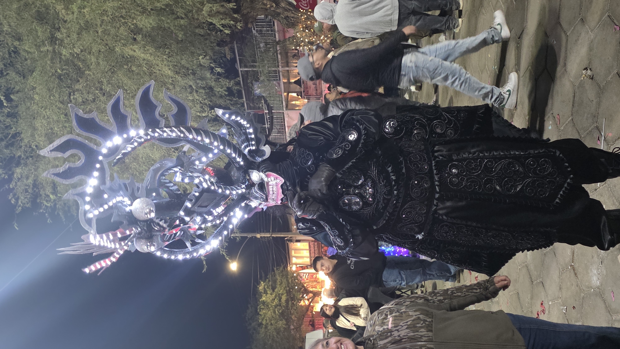 A person in a black, elaborate costume resembling a mythical creature or demon stands in the foreground of a nighttime street scene. The costume features a large, illuminated headpiece with horns and decorative elements, adorned with numerous white LED lights. The face of the mask is visible with pink teeth and white eyes. The body of the costume is also black and heavily embellished with intricate patterns and sparkling details. The individual wears black gloves.
The person is standing on a paved street, with other people milling about in the background. Several buildings and trees are visible. The atmosphere suggests an outdoor event or festival, possibly a parade or cultural celebration, given the festive lights and the decorated costume. The ground is littered with small pieces of confetti. In the far background, there are visible signs and lights from businesses, including a Coca-Cola advertisement. The lighting is a mix of the costume's bright LEDs, streetlights, and the ambient light from surrounding establishments, creating a vibrant nighttime ambiance. The location is identified as the city of Pozo Almonte, Chile.