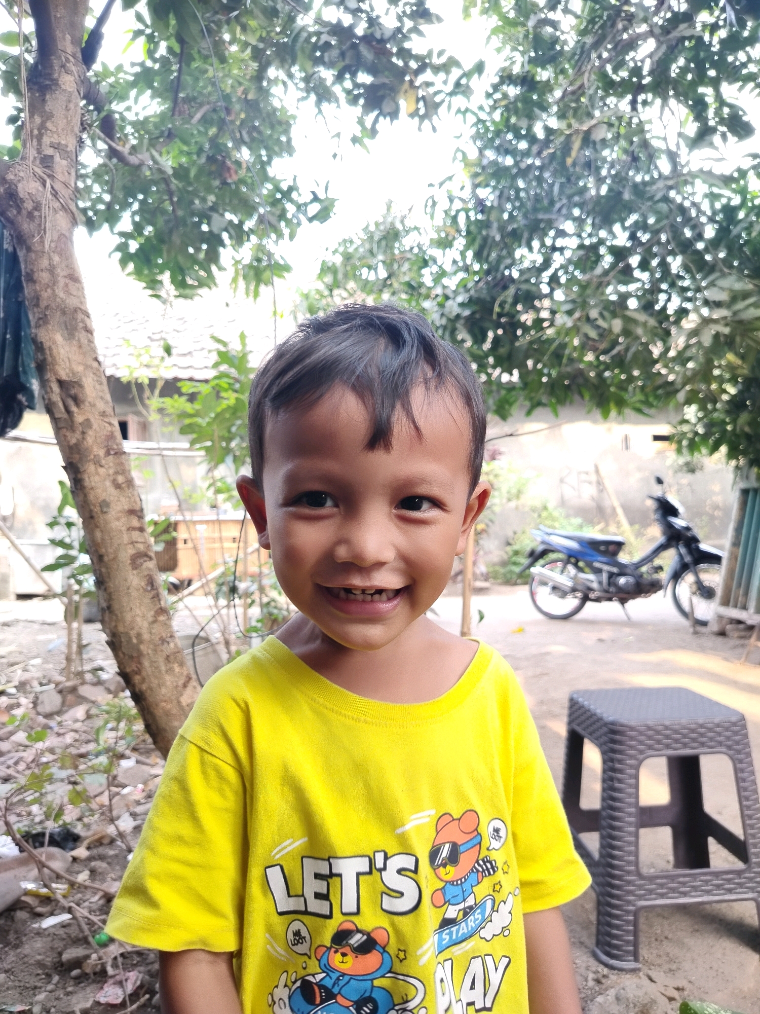 A young boy wearing a bright yellow t-shirt with graphics of cartoon characters is standing outdoors. The t-shirt has text that reads "LET'S" and "STARS PLAY" along with images of a bear wearing sunglasses and a blue jacket, and another bear character. The boy is smiling and looking towards the camera. In the background, there is a tree trunk to the left, a motorcycle parked on a dirt ground to the right, and some buildings and foliage. The scene appears to be in a residential or semi-rural area in Beloh, Indonesia. The lighting suggests it is daytime, possibly late afternoon given the dappled sunlight.
