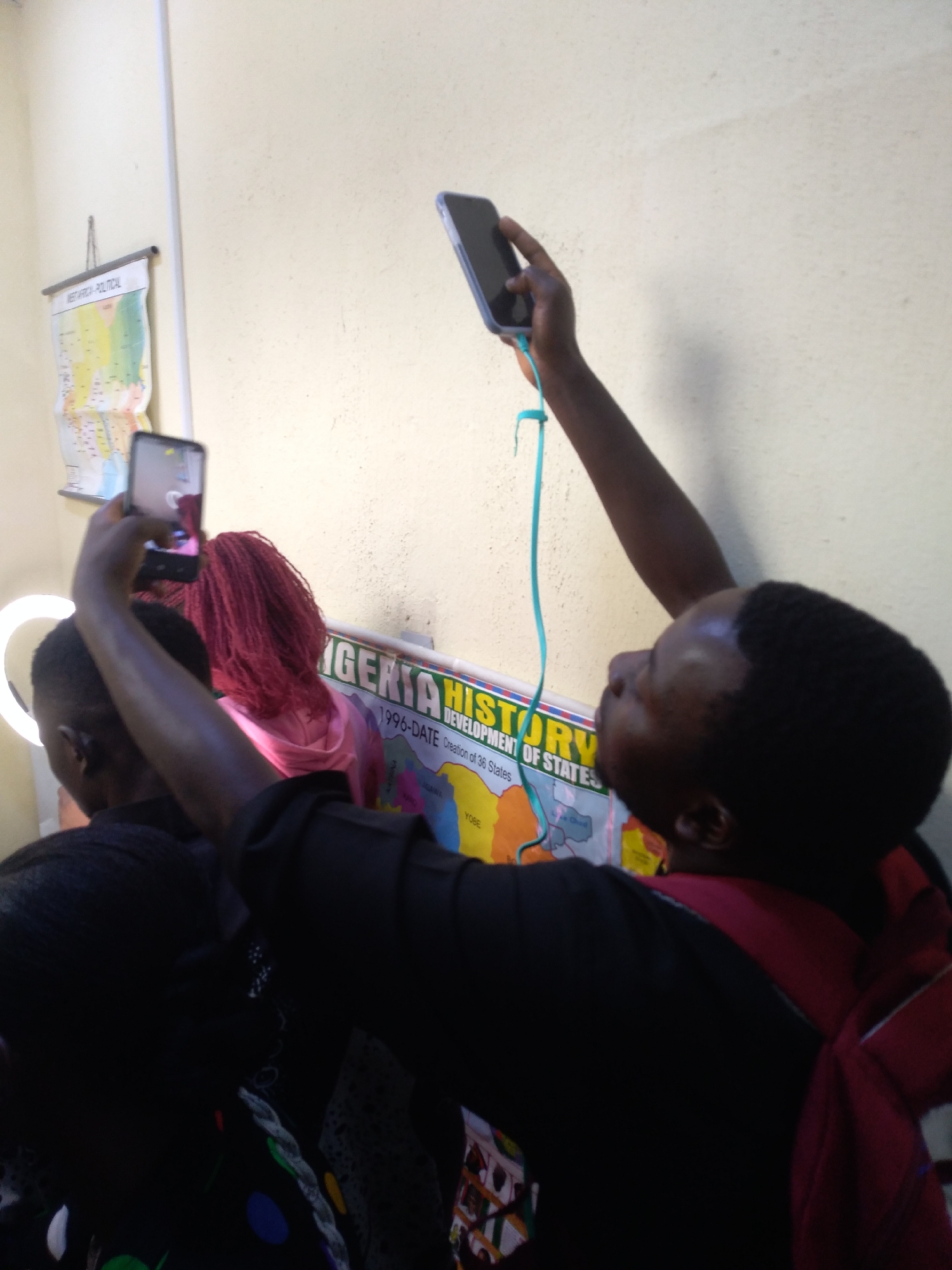 This indoor scene, set in Jos, Nigeria, shows several individuals engaged in recording or photographing an event. In the foreground on the right, a person with dark skin and wearing a dark shirt holds a smartphone high with their right hand, from which a blue lanyard hangs. Another individual, partially visible in the center-left, has distinctive red/pink hair and is also holding a smartphone up, its screen reflecting light. A third person is mostly obscured on the far left, positioned near a glowing ring light.
The setting appears to be an educational or presentation space, characterized by light-colored, textured walls. Two maps are displayed on the wall: one labeled "WEST AFRICA - POLITICAL," and a larger one titled "NIGERIA HISTORY." The history map further details the "DEVELOPMENT OF STATES" and specifies "1996-DATE Creation of 36 States," with "YOBE" and "GOMBE" visible among the state names. The activity suggests an audience or participants actively documenting a lecture, presentation, or performance, possibly for social media or educational purposes, given the ring light and multiple phones being used to capture content.