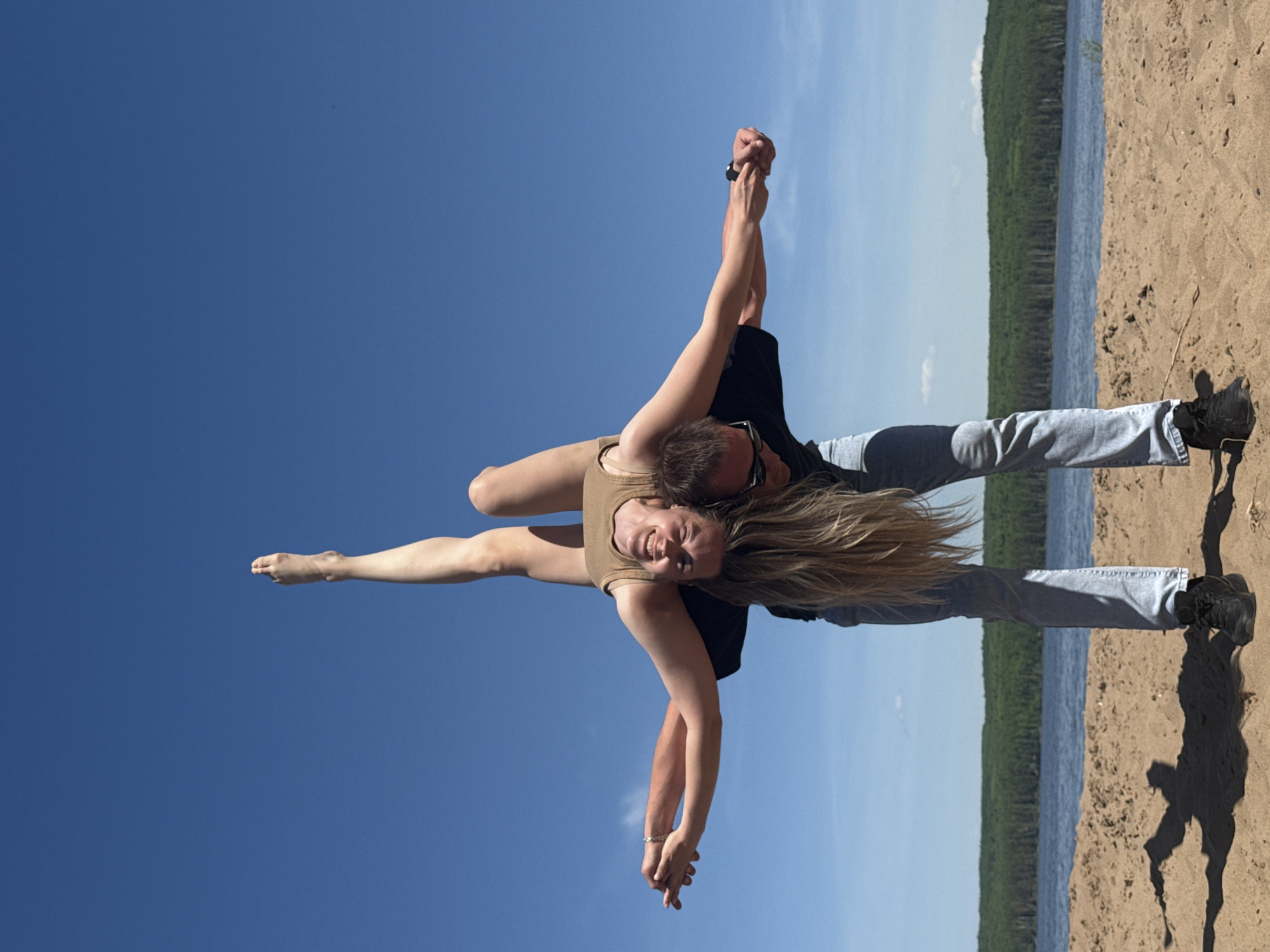 A man and a woman are performing an acrobatic move on a beach on a sunny day. The man is standing on the sand wearing jeans, a black shirt, and sunglasses. He is holding the woman in the air. The woman is wearing a tan-colored leotard and has her right leg extended straight up toward the blue sky. The beach is on the edge of a lake surrounded by trees. The image was taken in the city of Chaikovsky, Russia.
