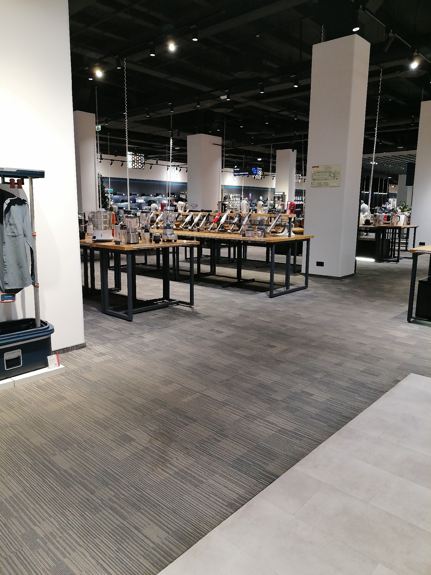 The image shows an interior view of a retail store, likely a department store or electronics store, in Rostov-on-Don, Russia. The store features rows of tables displaying kitchen appliances such as blenders and steamers. The ceiling is dark with multiple recessed lights. White structural columns are visible throughout the space. In the foreground left, a light blue garment is hung on a rack, and a dark blue container is at its base. The floor is carpeted with a grey, striped pattern. The overall impression is of a well-lit, organized retail environment.
