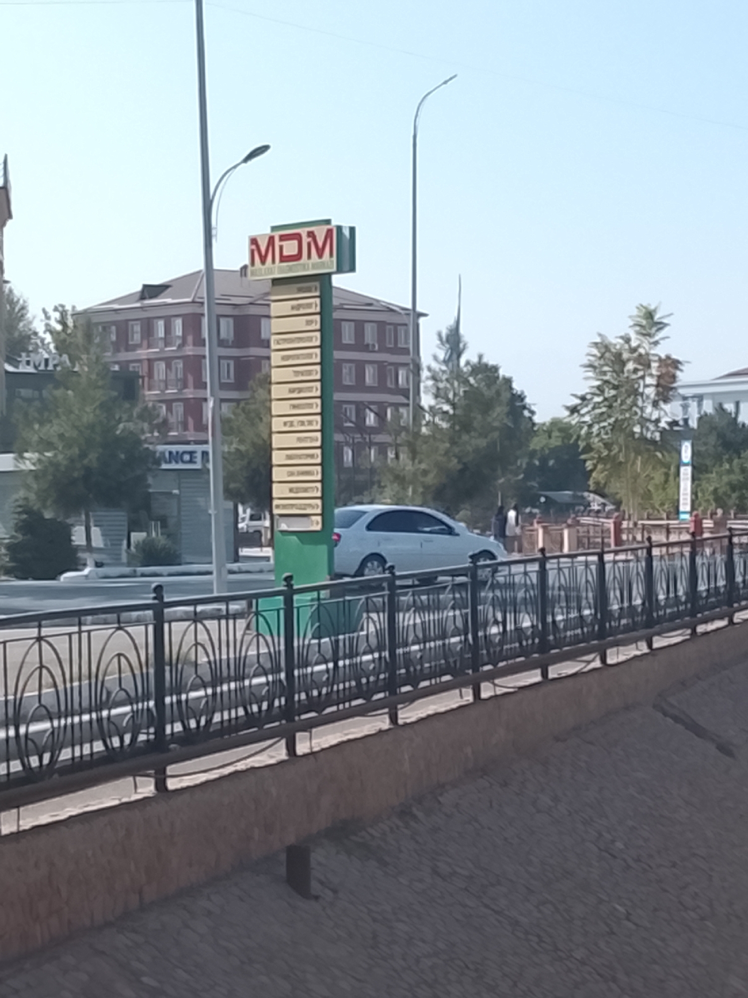 The image captures an outdoor scene in Fergana City, Uzbekistan, featuring a multi-story building with red brick facade and white window frames in the background. In the foreground, a decorative metal fence runs along a walkway or bridge. A light-colored sedan is parked on the road next to the walkway. A tall signpost with the letters "MDM" in red and white is prominently displayed on the left side of the image. Below "MDM," a list of smaller text entries is visible on the sign, suggesting it might be directory or advertisement. Trees and streetlights are also present in the scene, contributing to the urban environment. The sky is clear and bright, indicating a daytime setting.
