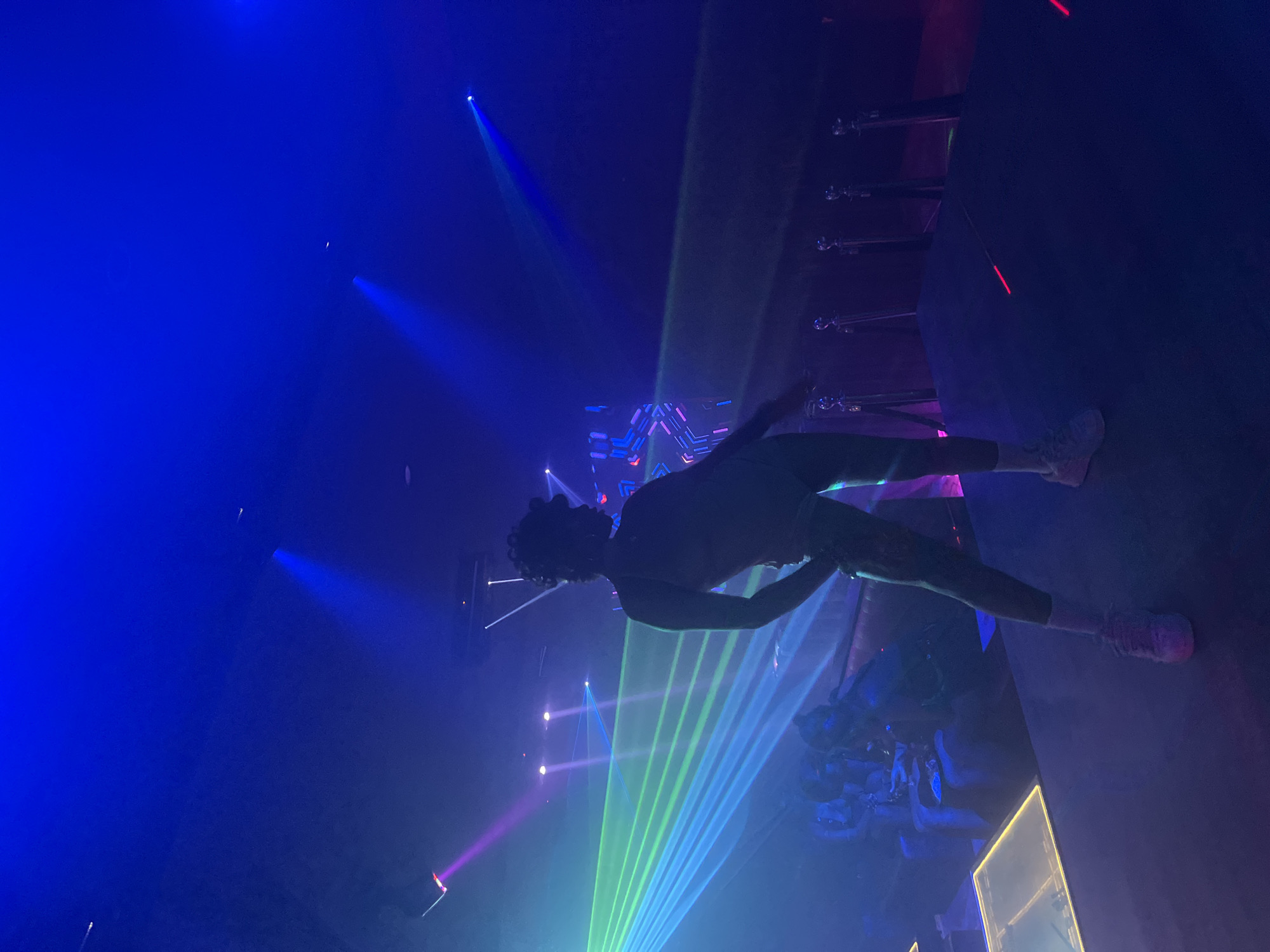 The image depicts a person standing on a stage, silhouetted against a backdrop of vibrant blue and green laser lights. The individual appears to be performing, with their body angled and one arm extended. The stage is dark, with a few red lights visible on the floor. In the background, other individuals can be seen seated at tables, and the environment suggests a nightclub or entertainment venue. The overall atmosphere is dynamic and energetic, characterized by the dramatic lighting and the focus on the performer. The location is specified as Howayi, Nigeria, implying an urban setting within that region.