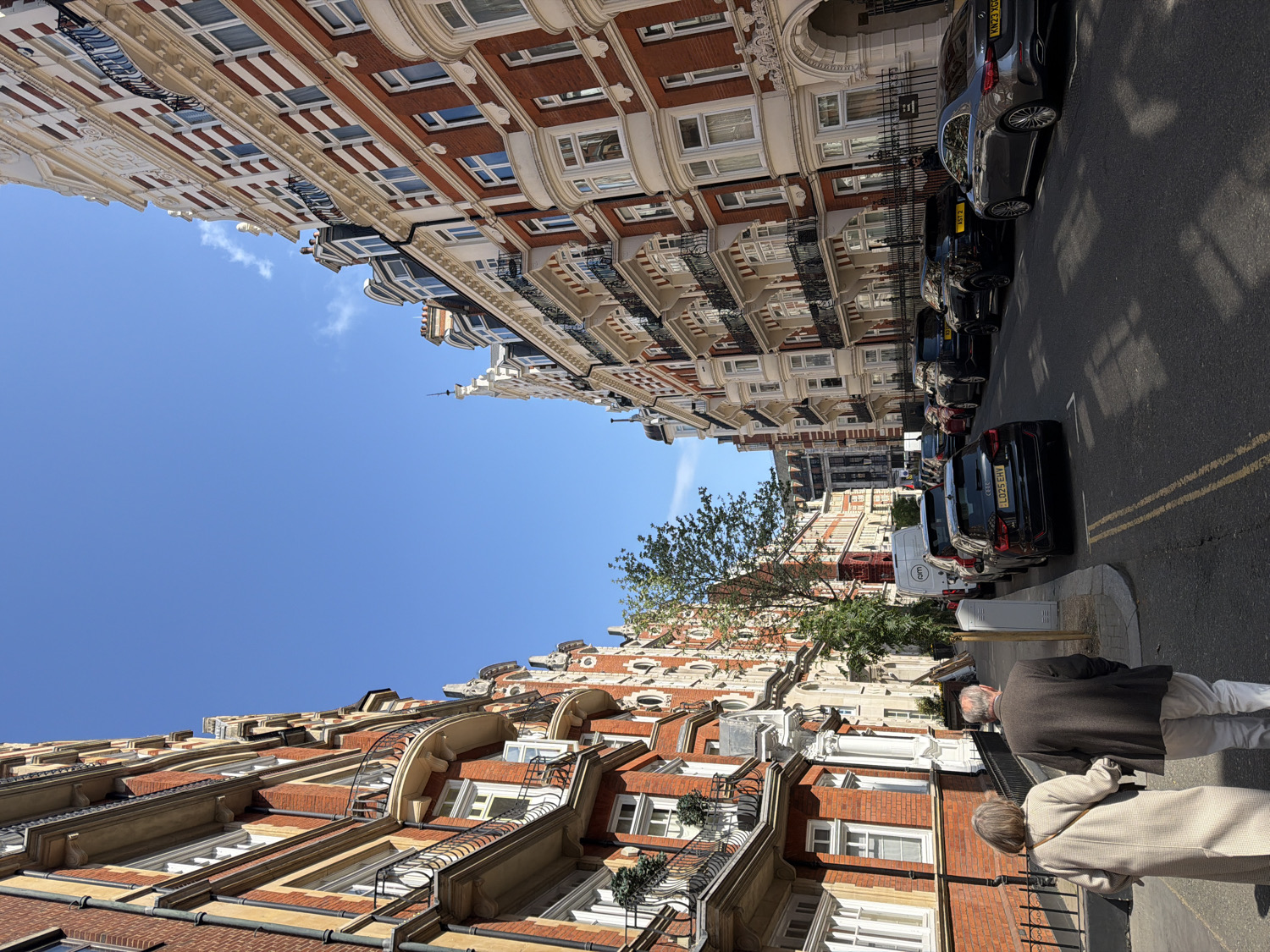 The image displays a street scene in an affluent urban area of London, United Kingdom, under a clear, bright blue sky with a few wisps of white clouds. The perspective looks along a street lined on both sides by grand, multi-story buildings.
These buildings are architecturally distinctive, featuring red brickwork contrasted with elaborate cream or white stone facades. They are adorned with ornate details such as projecting bay windows, curved balconies with black wrought-iron railings, decorative cornices, and classical pilasters, suggesting a period of significant architectural opulence.
On the street level, a row of cars is parked along the curb on the right side. Several UK license plates are visible, including "KV23 XGU," "LD23 EHY" on black sedans, and "KVV7 CAA" on another vehicle. A white commercial van with the "cmt" logo and license plate "L1025 EHY" is also parked. The road surface has distinctive yellow double lines.
In the foreground on the left, two individuals are seen from behind, walking along the sidewalk. One is an older man with grey hair, wearing a dark jacket, and the other is an older woman with light-colored hair, dressed in a light-colored coat. Green trees are interspersed along the sidewalk. The strong sunlight casts clear shadows, indicating it is a bright day, likely midday or early afternoon. The scene conveys a sense of serene urban elegance.