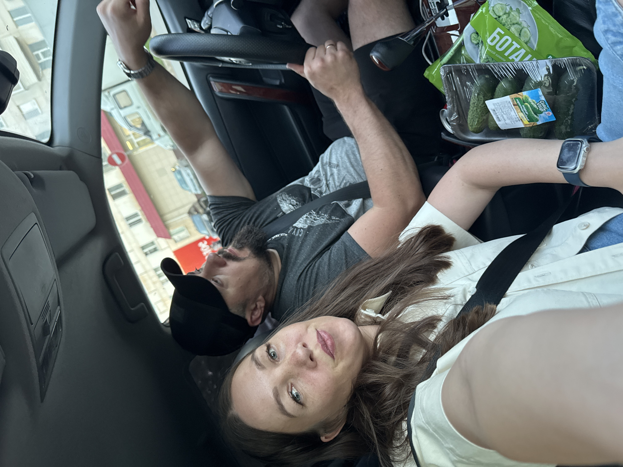 A selfie taken inside a vehicle, with two individuals. The woman, seated in the passenger seat, is positioned at the bottom center of the frame, looking directly at the camera. She has brown hair and is wearing a white top and a seatbelt. Her left arm is visible, adorned with an Apple Watch. Next to her, a container of cucumbers with the label "Global Village" rests. Another package of cucumbers labeled "БOTAK" is visible behind it. A man is seated behind the steering wheel, wearing a black cap and a grey t-shirt. He has his arm raised near the roof of the car. The background shows urban surroundings, with buildings, including one featuring a red sign for "JOE," and a street. The photograph was taken in the city of Chaikovsky, Russia.