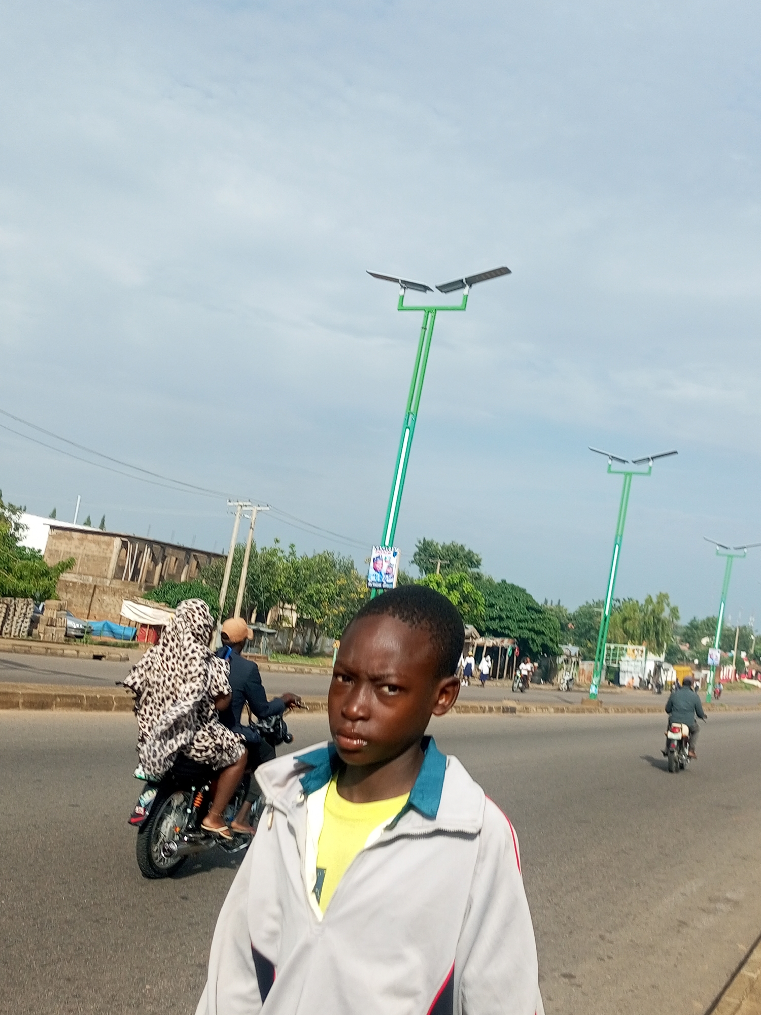 In Tempure, Nigeria, a boy in a grey jacket with a yellow shirt peeks into the frame from the right, looking directly at the camera with a serious expression. Behind him, a motorcycle carrying a person in a leopard-print garment travels on a paved road under a partly cloudy sky. In the background, modern streetlights with solar panels stand tall along the roadside, bordering buildings and lush green trees, suggesting a developing urban environment. The scene depicts a typical day on the streets of Tempure, with everyday activities and infrastructure coexisting.