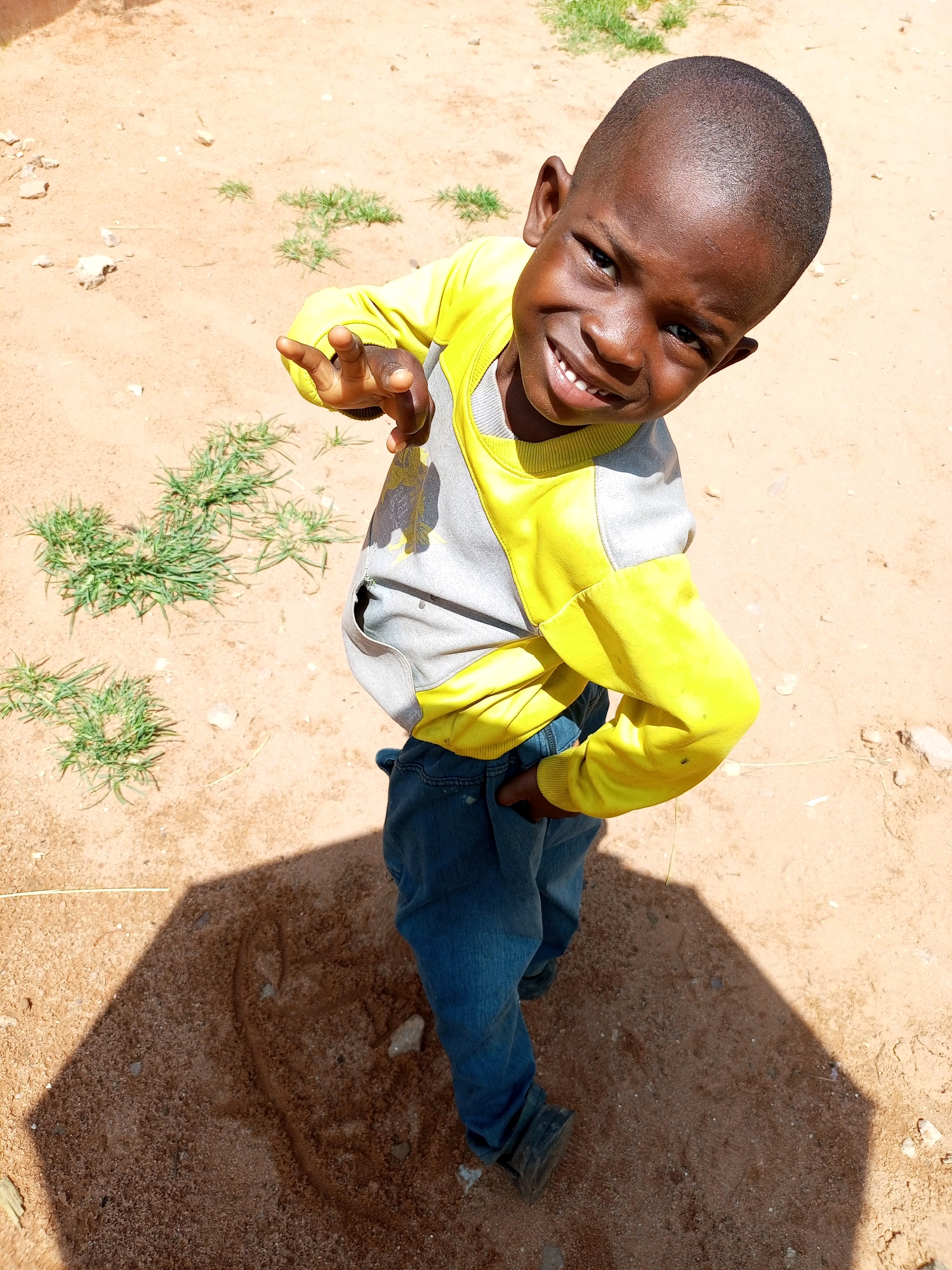 A high-angle medium shot captures a young Black boy standing on sandy ground in Tempure, Nigeria. He is smiling broadly and looking directly at the camera, with his head tilted to the right. The boy is wearing a yellow and gray long-sleeved shirt and blue jeans. His right hand is raised, with his index and middle fingers extended, as if he is gesturing or making a peace sign. The ground is dry and dusty, with patches of sparse green grass and a few scattered rocks. A distinct hexagonal shadow, cast by an unseen object, covers a portion of the ground to the boy's left. The lighting suggests it is daytime, likely with strong sunlight.