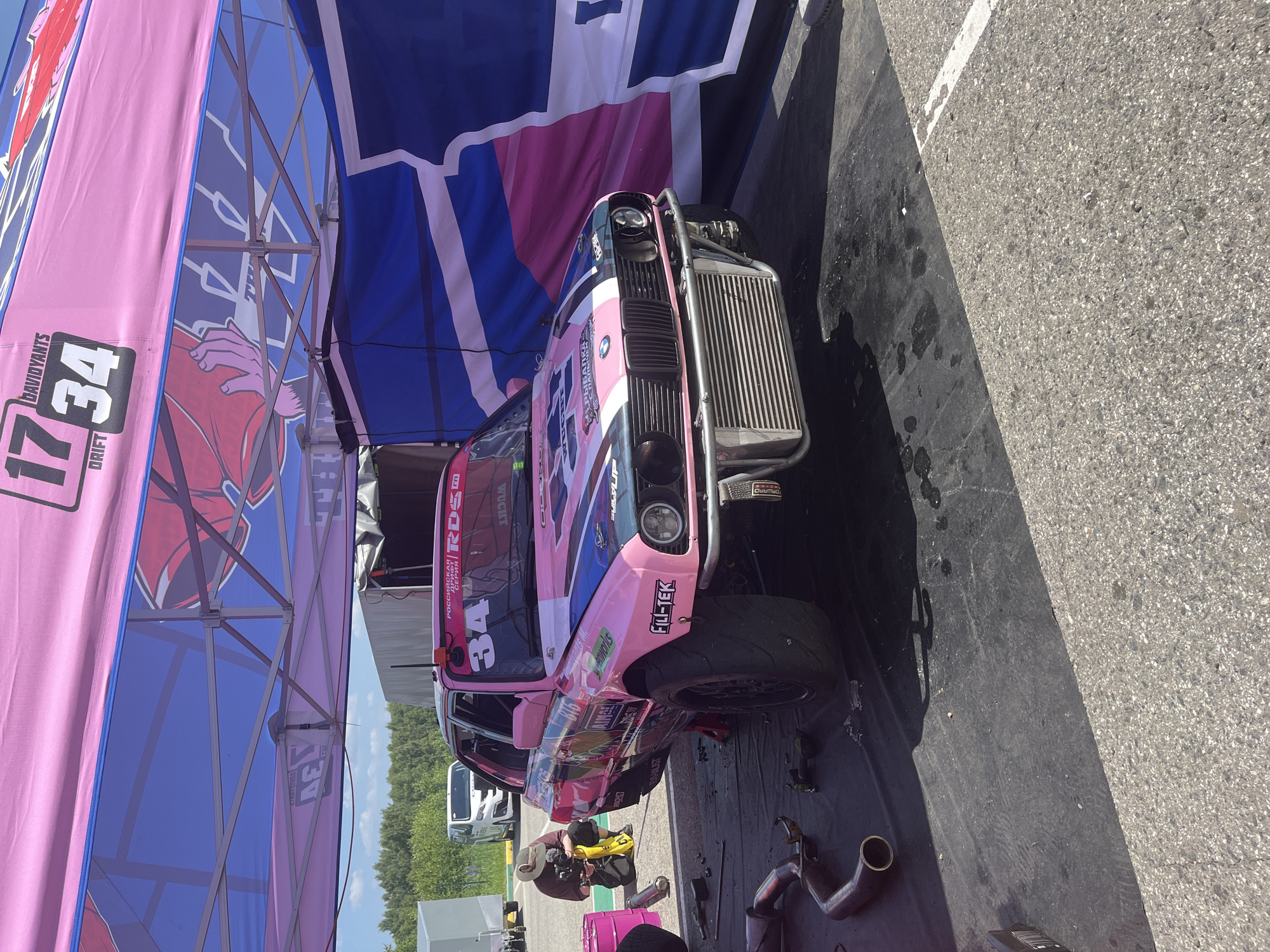 The image captures a pink drift car, identified by the number "34" and various sponsor logos, parked under a large pink and blue tent structure. The car has a custom front bumper with a large intercooler visible. The scene appears to be a pit area, as indicated by tools and exhaust pipes scattered on the black matting in front of the car. A person wearing a hat and a dark shirt is kneeling behind the car, operating a camera. In the background, a white van and trees are visible, suggesting an outdoor location. The tent is adorned with branding, including "DAVIDYANTS DRIFT" and "RDS", potentially indicating a drift racing event. The ground transitions from the black matting to asphalt. The overall environment suggests a racing or automotive event setting in the city of Beryozovka, Russia.