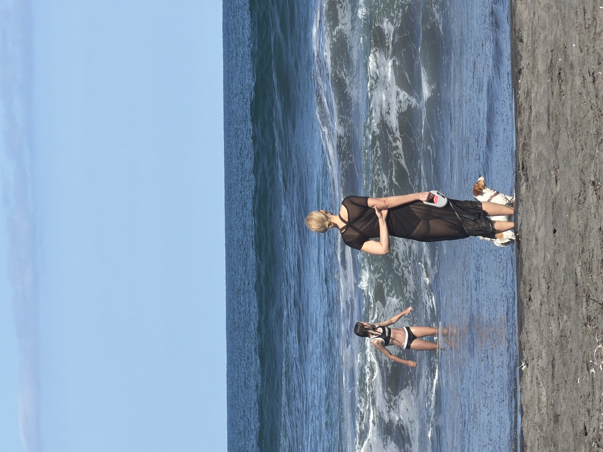 This is a vertical, full-shot photograph taken at what appears to be a beach in Yuzhno-Kurilsk, Russia. The scene captures two individuals and a dog. In the foreground, a young person in a black and white bikini stands in the shallow ocean water, their arms outstretched. Behind them, a woman with blonde hair tied up, wearing a sheer black dress over dark swimwear, stands on the wet sand near the waterline. She holds a light-colored object, possibly a small bag or pouch. A small dog with brown and white markings stands next to her, facing away from the camera. The ocean is calm with small waves and foam breaking on the shore, indicating a gentle tide. The sky is a clear, light blue, suggesting it is daytime with pleasant weather. The sand is dark and wet from the receding water. The overall mood of the image is relaxed and leisurely, suggesting a day spent at the beach. There is no visible text in the image.