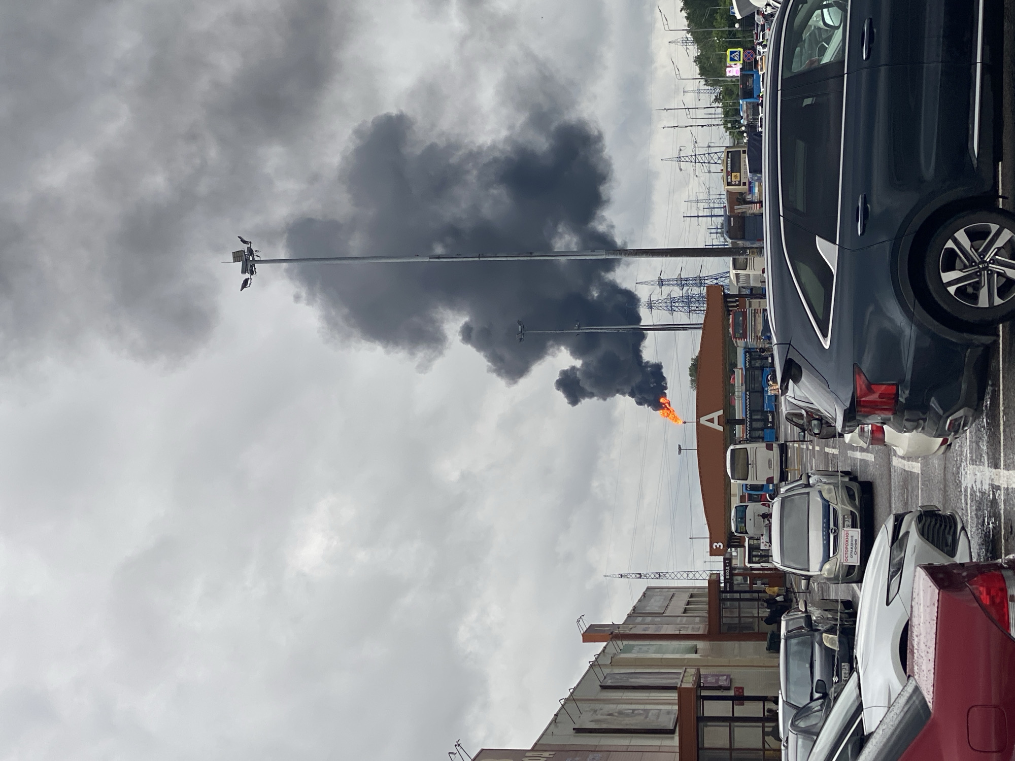 The image depicts a commercial or industrial area in Moscow, Russia, with a large plume of dark smoke rising into an overcast sky, suggesting a fire or industrial process. In the foreground, a parking lot is filled with numerous cars, including a dark SUV and a white sedan. In the mid-ground, several buses are parked, and people can be seen walking around the buildings. One building, with signage that includes the letter "A," appears to be a retail or transportation hub. In the background, numerous power lines and towers are visible, indicating a developed urban or industrial landscape. A flare or controlled burn with orange flames is visible at the base of the smoke plume. The overall scene conveys a sense of an active event that is producing significant smoke.