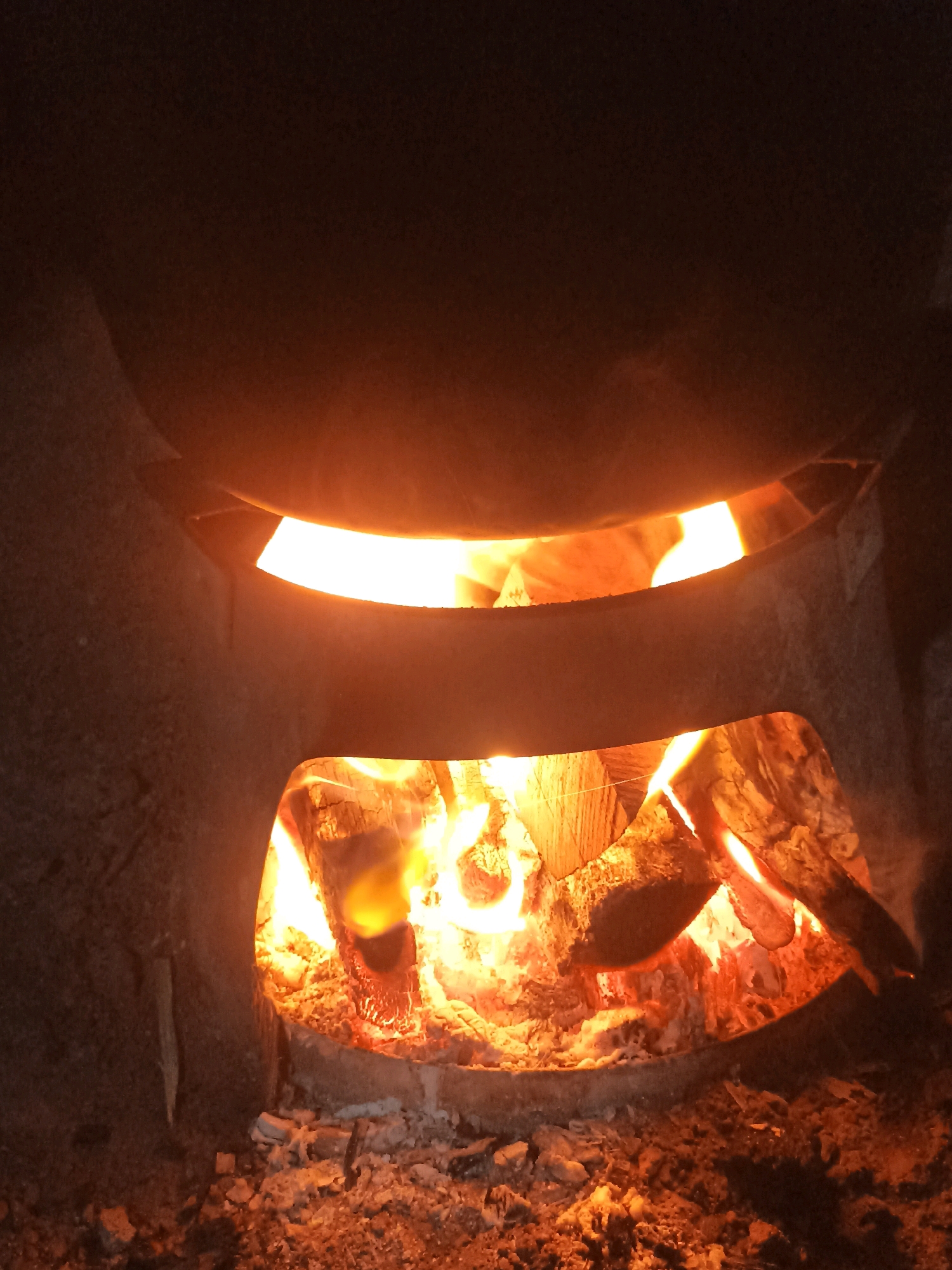 The image shows a close-up view of a wood-burning stove in Mojopanggung, Indonesia. The stove is made of dark, rough material and has two openings. The lower opening reveals a fire burning with bright orange and yellow flames. Several pieces of wood are visible, some partially consumed by the fire, resting on a bed of glowing embers and ash. The upper opening shows a glowing orange light, suggesting more burning material or heat within. The overall scene is dimly lit, with the primary illumination coming from the fire itself. The background is dark and out of focus.