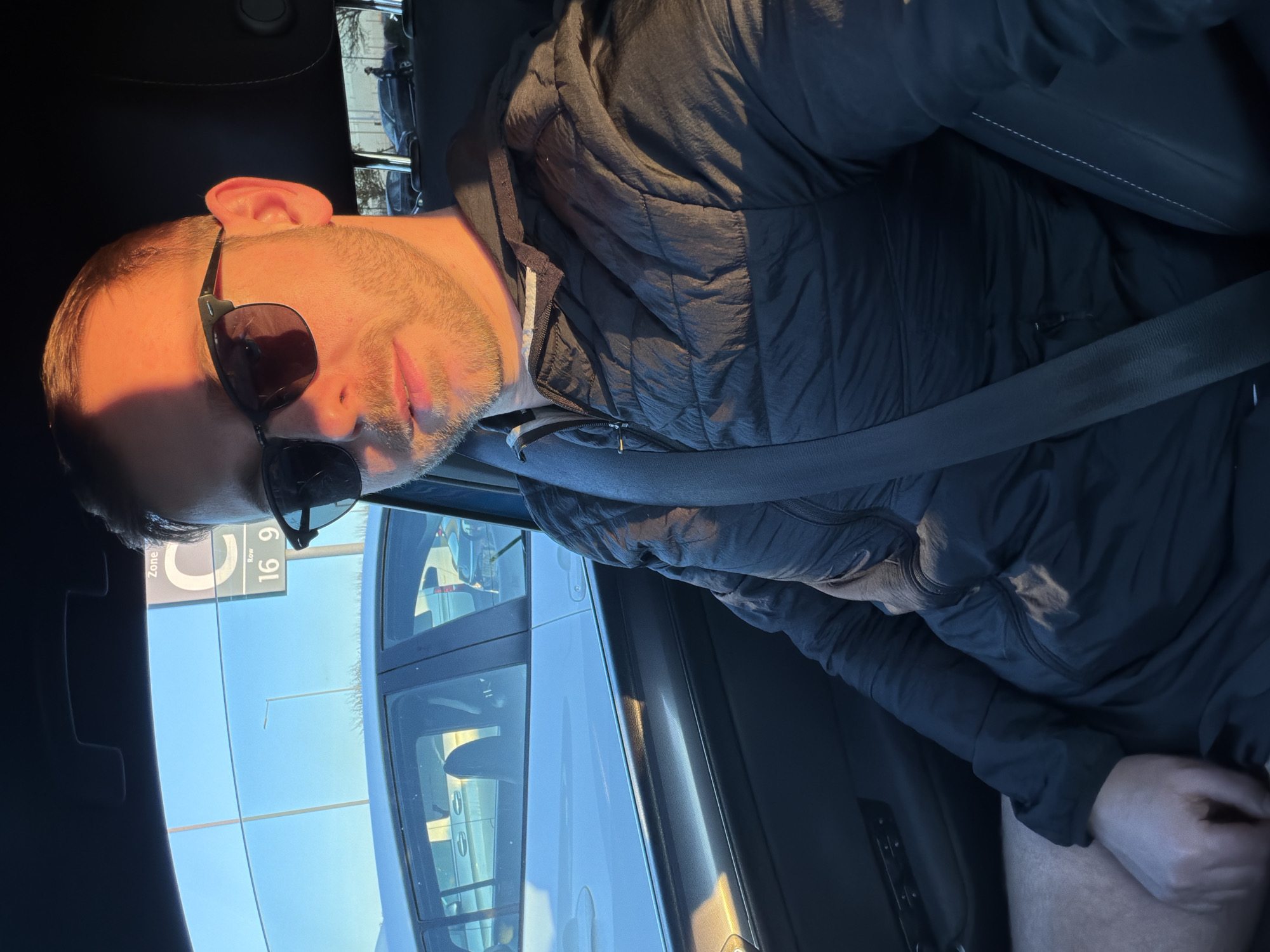 A man is sitting in a car, wearing sunglasses and a black jacket, with a seatbelt fastened. The car is parked in a parking lot labeled "Zone C, Row 16". The location appears to be London, United Kingdom. The day is sunny, with clear skies.
