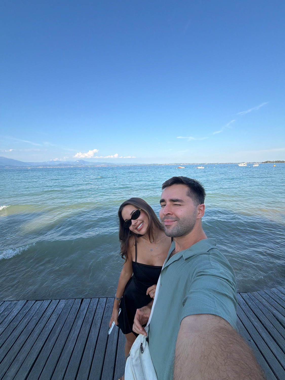 A man and a woman stand on a wooden pier overlooking a body of water under a clear blue sky. The man, on the right, wears a light green collared shirt and takes the selfie, winking at the camera. The woman, to his left, wears a black dress and sunglasses, smiling. In the background, boats dot the water, and distant mountains are visible. The scene is consistent with the setting at the Sirmione Peninsula in Italy, known for its scenic lake views and historic architecture.