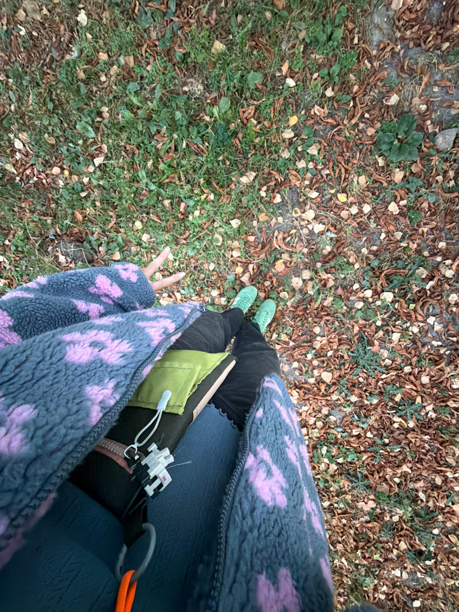 The image is a first-person perspective shot looking down at a person's legs and torso, with a ground covered in grass and fallen leaves. The person is wearing a grey fleece jacket with purple tie-dye patterns, dark pants, and bright green sneakers. A green pouch is attached to the jacket's zipper, with a keychain featuring a white and black object. The person is holding up two fingers in a peace sign with their right hand. The environment suggests an outdoor setting, possibly a park or a wooded area, with fallen leaves indicating autumn. Given the overall context and style of the image, it is likely taken in a city like Kyiv, Ukraine, perhaps during a walk in a public space.