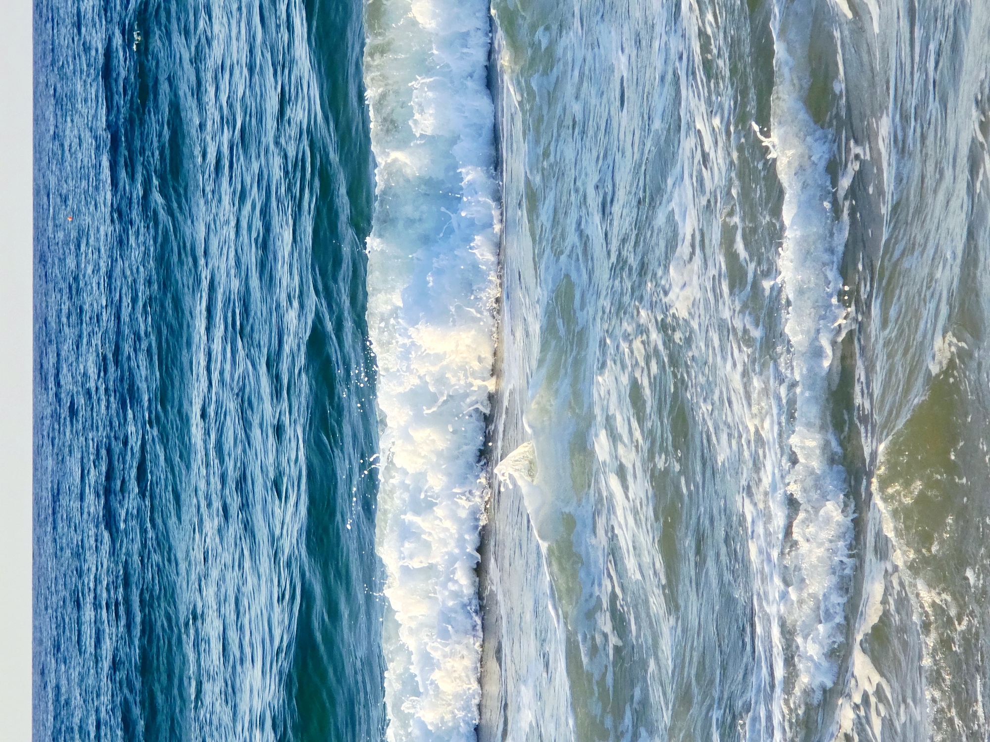 The image depicts a close-up view of the Aegean Sea, with waves rolling towards the shore. The water is a vibrant blue, transitioning to a lighter turquoise and white near the breaking waves. The sky is a pale, hazy white, indicating it is either early morning or late evening, or a cloudy day. The ocean surface is dynamic, with visible ripples and foam on the crests of the waves. There are no people or man-made structures visible in this frame, only the natural elements of the sea. This scene is consistent with the coastline of Malia Municipal Unit, Greece, known for its beaches and clear waters.