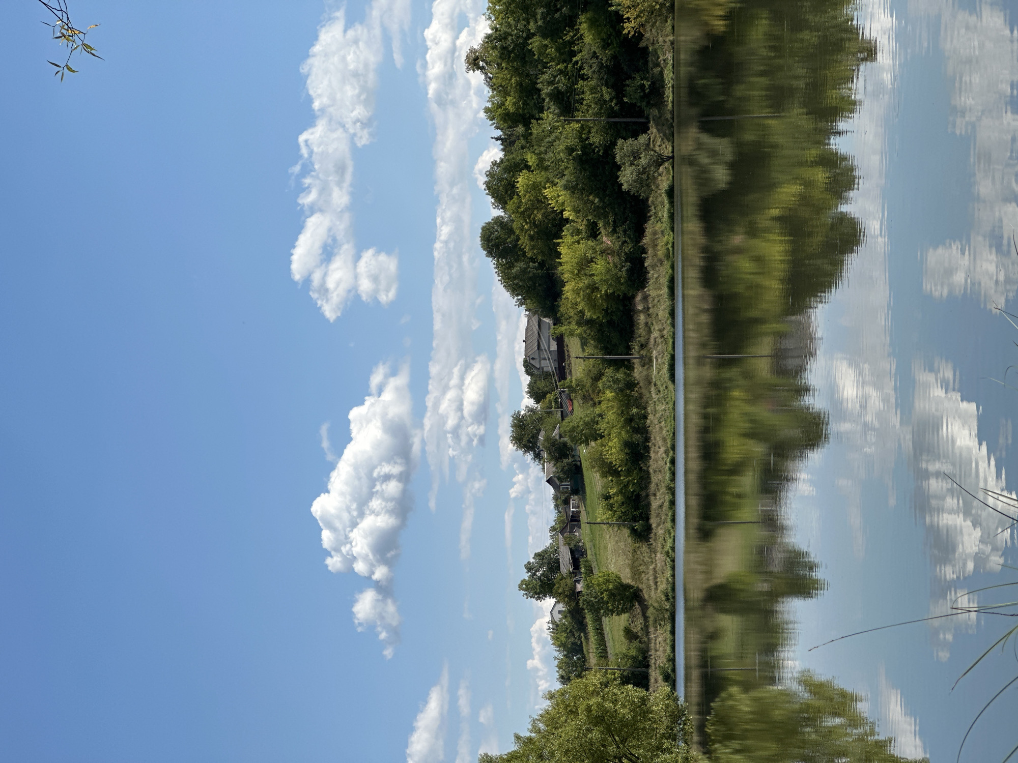 The image depicts a tranquil lakeside scene on a clear, sunny day with scattered white clouds in a bright blue sky. The foreground is dominated by a calm body of water, reflecting the sky and the lush green foliage of the opposite bank. The embankment is lined with trees and vegetation, and further inland, a small cluster of houses is visible nestled on a gentle slope. A few power poles are also present along the embankment. The overall atmosphere is serene and natural, suggesting a rural or semi-rural setting, consistent with the village of Makiv, Ukraine. There are no people or animals visible in the image.