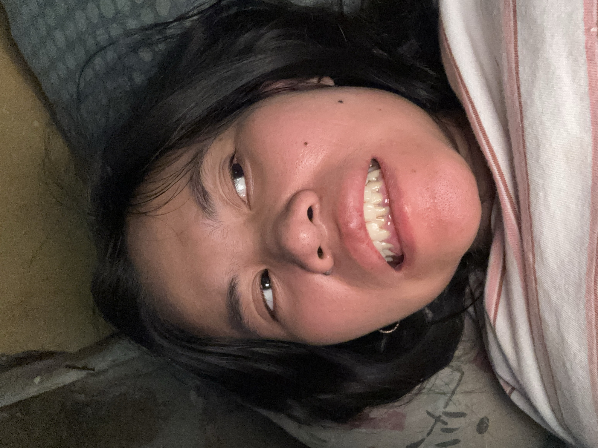 A close-up, low-angle, indoor portrait captures a young woman with dark hair lying down, her head tilted and eyes looking off to the right. She is wearing a striped shirt. The background is dimly lit and appears to be a bed with a patterned blanket and a dark, textured surface. The woman's expression is ambiguous, with her mouth slightly open, revealing her teeth. The lighting is artificial and casts shadows, suggesting the photograph was taken at night. There is no indication of the specific location within East Jakarta, Indonesia.